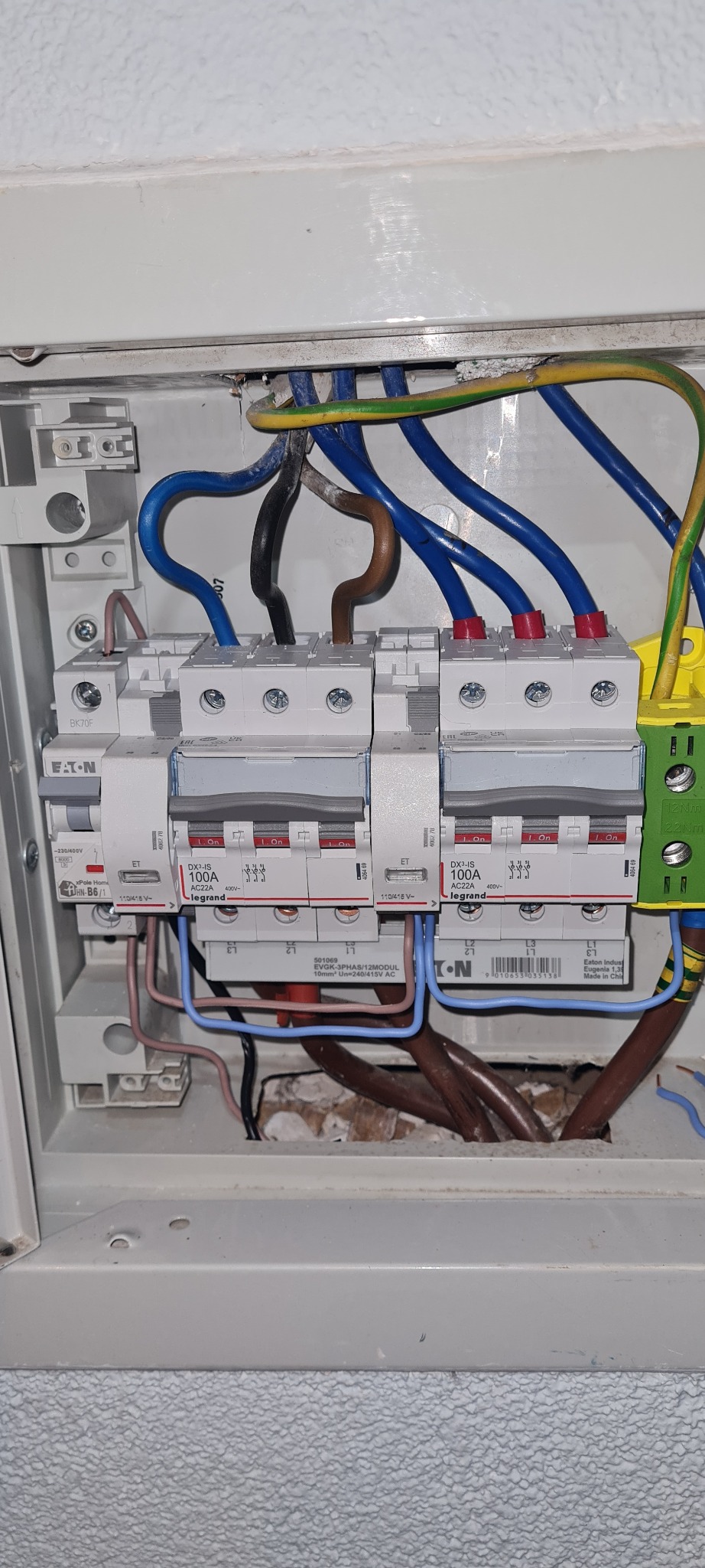 Otwarta skrzynka elektryczna z widocznymi bezpiecznikami, przewodami w kolorach niebieskim, brązowym, czarnym i żółto-zielonym, oraz elementami firm Eaton i Legrand, zamontowana na szarej ścianie...