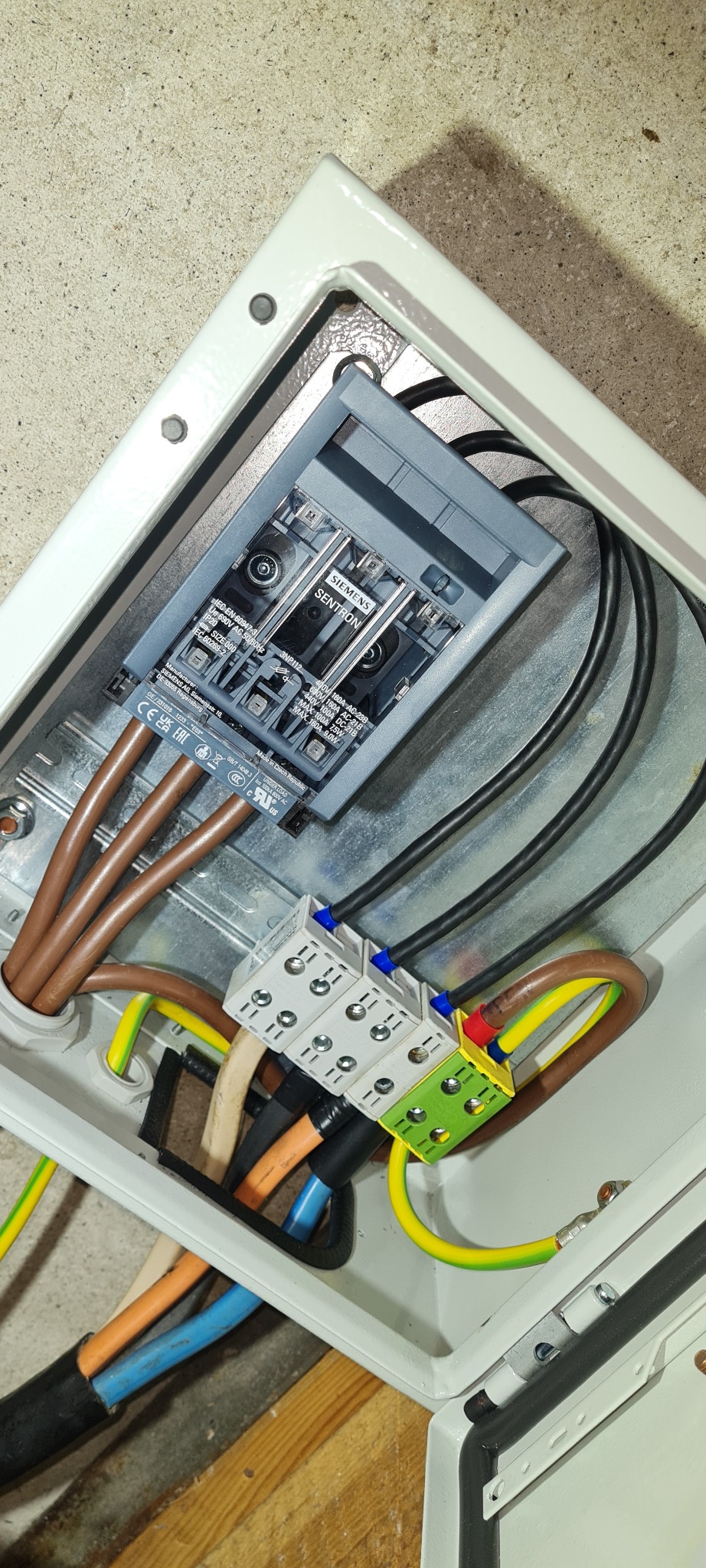 Rozdzielnica elektryczna w białej obudowie z zamontowanym bezpiecznikiem SENTRON firmy Siemens i podłączonymi przewodami o różnych kolorach (brązowe, czarne, niebieskie, żółto-zielone) do listwy...