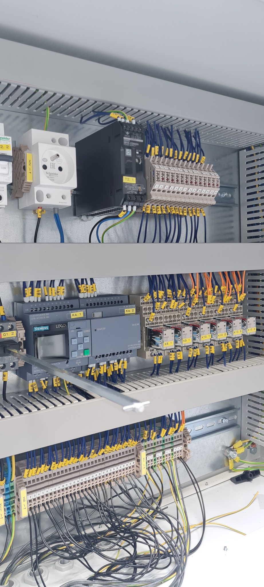 Wnętrze rozdzielnicy elektrycznej z modułem sterującym Siemens LOGO!, widoczne liczne przewody oznaczone żółtymi numerkami, listwy zaciskowe i moduły Schneider Electric, całość w metalowej obudowie.