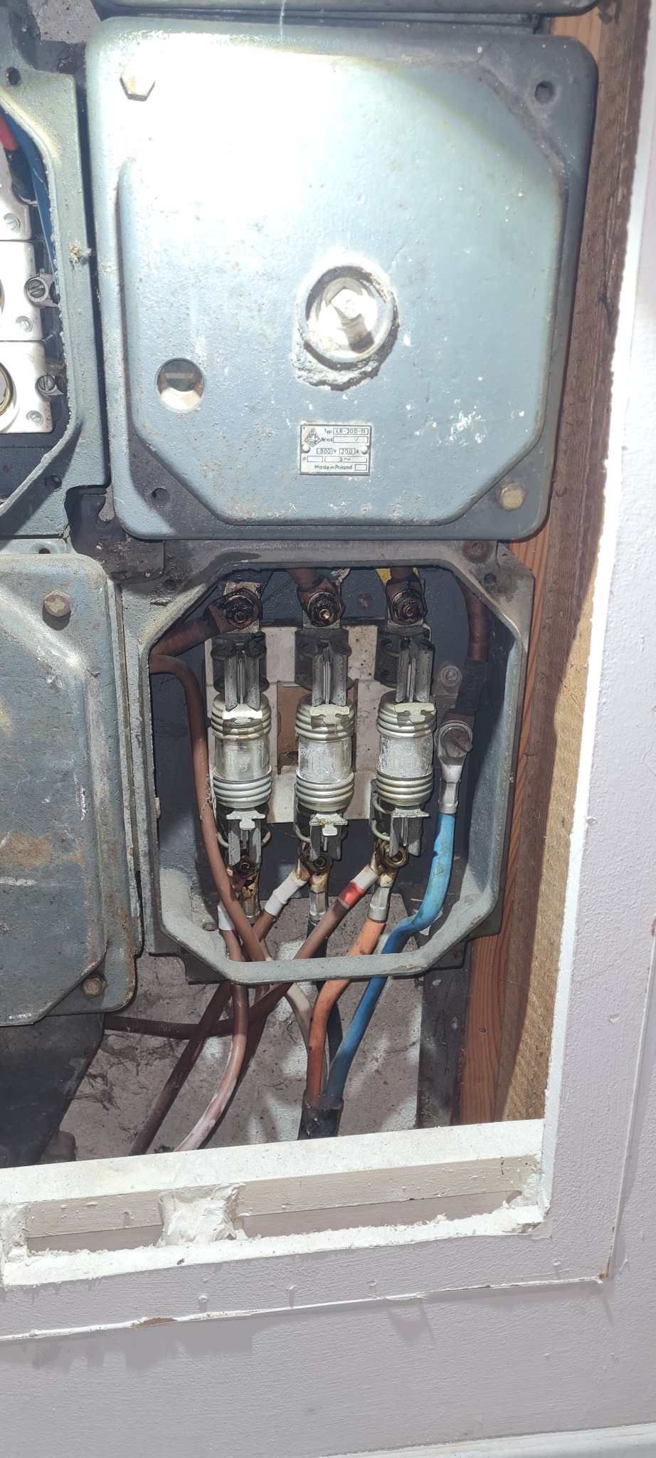 Otwarta, stara, szara skrzynka elektryczna z trzema bezpiecznikami topikowymi, widoczne przewody w izolacji brązowej, niebieskiej i pomarańczowej.
