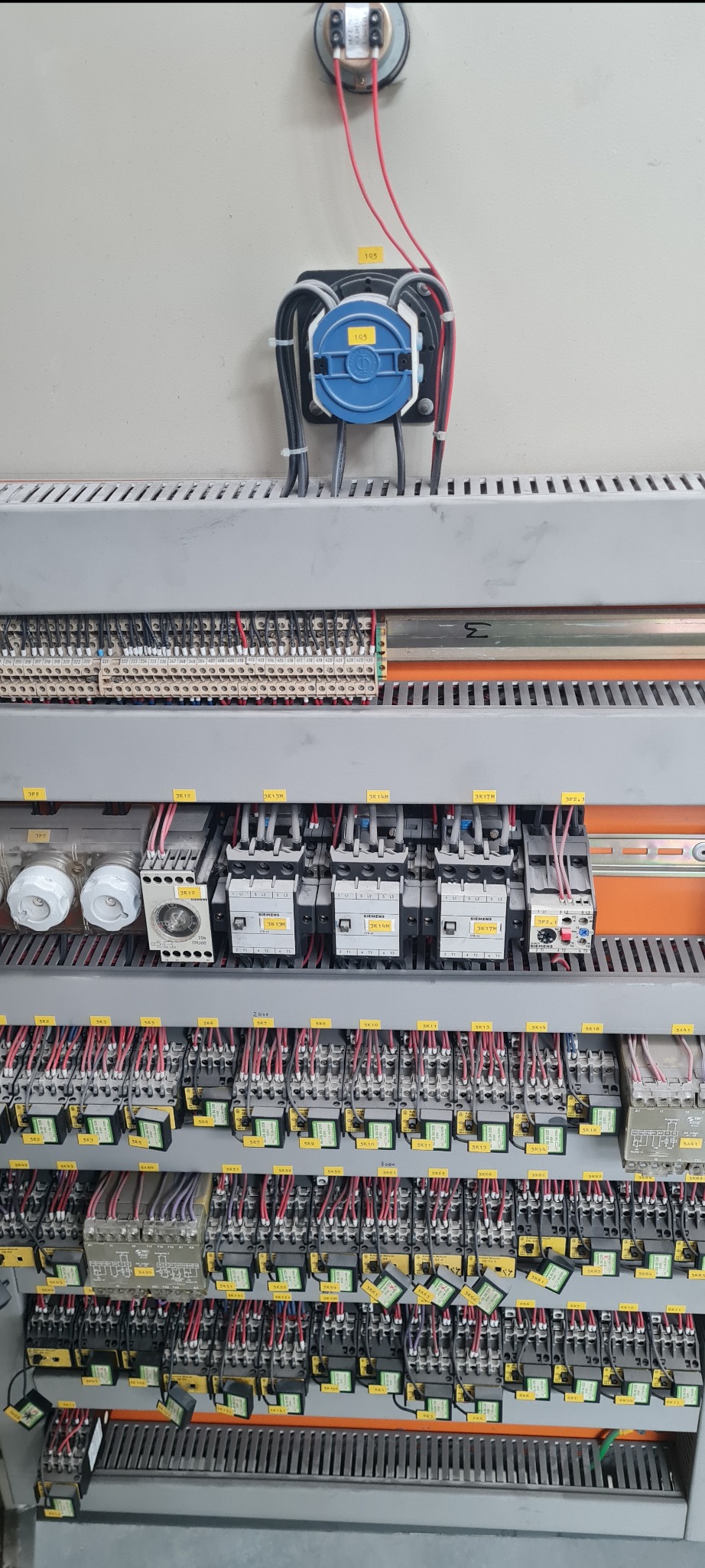 Rozbudowana szafa sterownicza z licznymi przekaźnikami, bezpiecznikami i okablowaniem oznaczonym żółtymi etykietami, widok z bliska na elementy wewnątrz szafy.