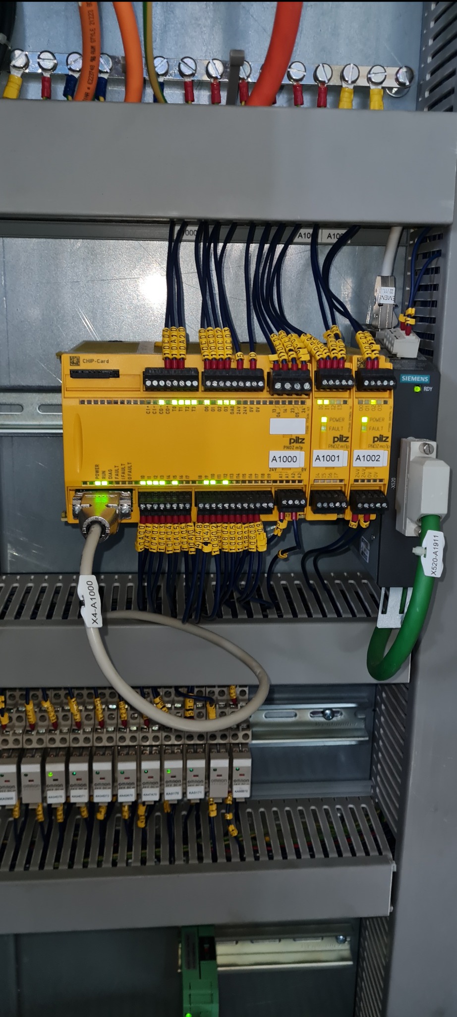 Rozdzielnica elektryczna z modułami bezpieczeństwa Pilz PNOZmulti, okablowaniem oznaczonym żółtymi tulejkami i listwami zaciskowymi, widoczne diody LED sygnalizujące stan pracy, w tle szyna montażowa.