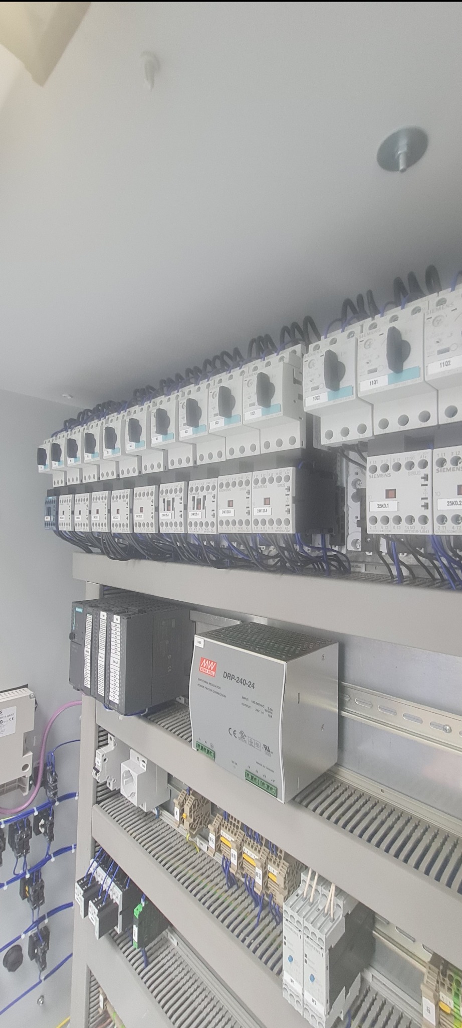 Rozdzielnica elektryczna z zainstalowanymi bezpiecznikami, stycznikami, zasilaczem impulsowym Mean Well DRP-240-24 i modułami sterującymi Siemens, widoczne liczne przewody w kolorze niebieskim...