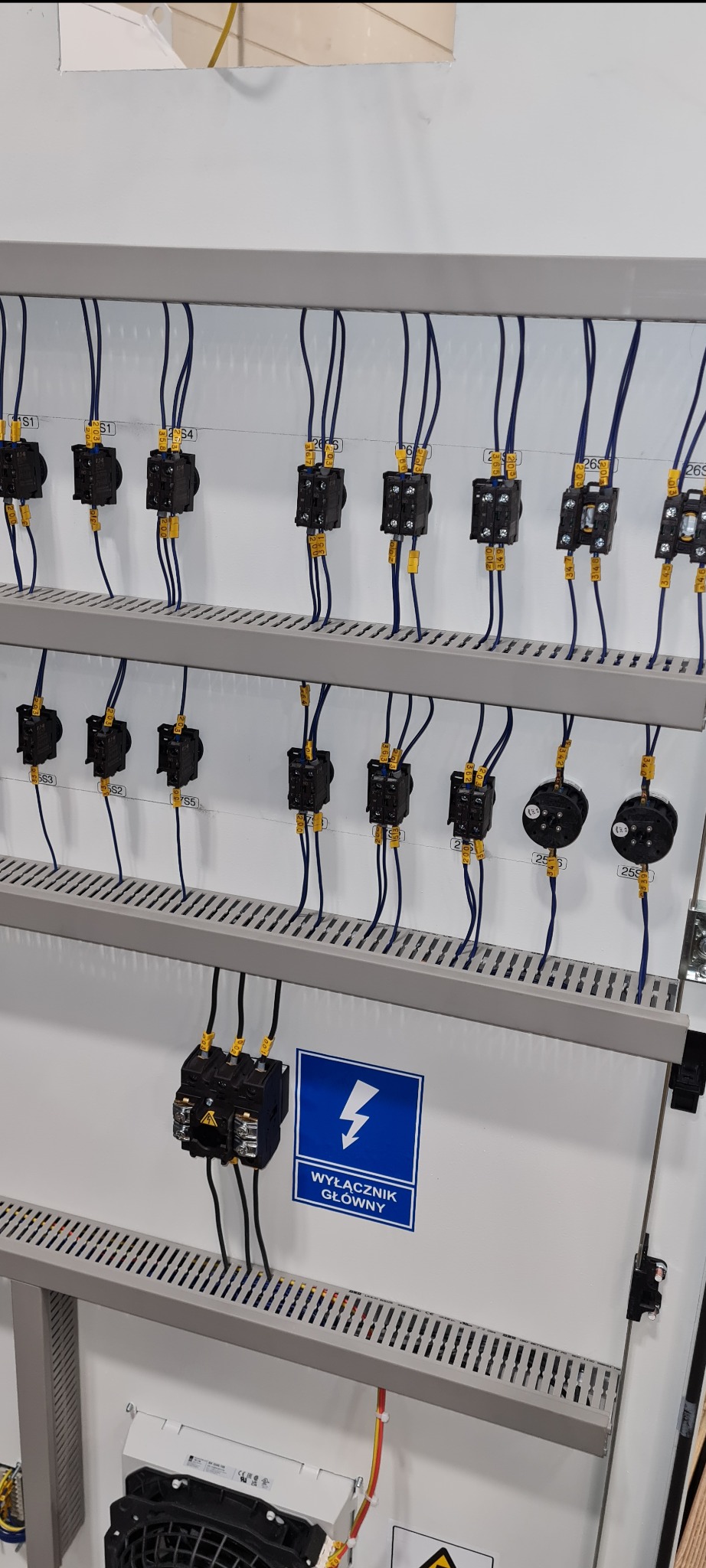 Rozdzielnica elektryczna z widocznymi bezpiecznikami, okablowaniem oznaczonym numerami i tabliczką ostrzegawczą 'Wyłącznik główny'.