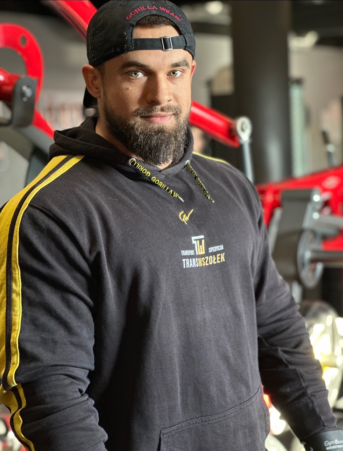Mężczyzna z brodą w czarnej bluzie z kapturem i czapce z daszkiem, pozujący na tle czerwonych maszyn na siłowni.