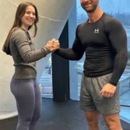 Uśmiechnięty trener personalny i klientka ściskają sobie dłonie w nowoczesnej siłowni, okno z widokiem na miasto w tle, sportowa atmosfera.