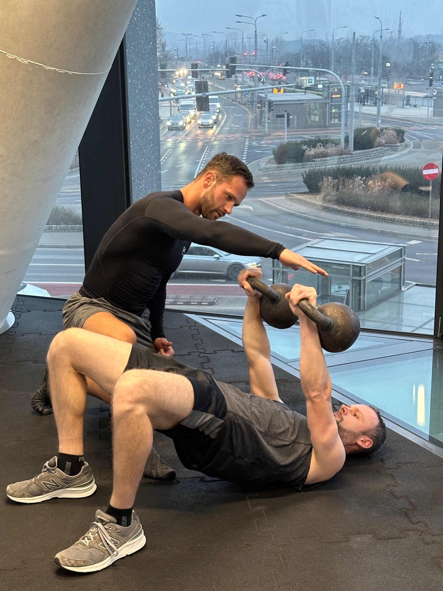 Mężczyzna ćwiczy z kettlem pod nadzorem trenera w siłowni z widokiem na miasto. Kettlebell trzymany nad głową w leżeniu na plecach, podnoszenie bioder.