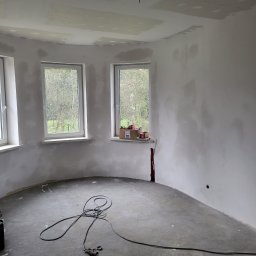 ANDROBUD - Wnętrze okrągłego pokoju w trakcie szpachlowania ścian i sufitu. Widoczne trzy okna, szpachlowane ściany, sufit z płyt kartonowo-gipsowych i kable na podłodze.