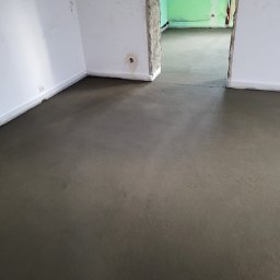 ANDROBUD - Wylana, świeża wylewka podłogowa w jasnym pomieszczeniu z białymi ścianami. Widoczne ślady po instalacjach w ścianie. W tle przejście do kolejnego pomieszczenia.