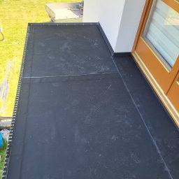 ANDROBUD - Wykończony balkon z czarną, hydroizolacyjną powłoką, widoczne ślady stóp. Detal wykończenia przy drzwiach balkonowych i metalowe obrzeże. Trawa w tle.