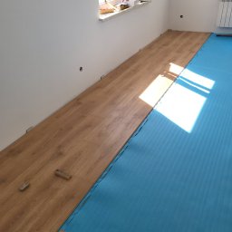 ANDROBUD - Układanie paneli podłogowych w jasnym wnętrzu. Częściowo ułożona podłoga z paneli w odcieniach drewna, podkład z niebieskiej pianki. Widoczne narzędzia i fragment okna.