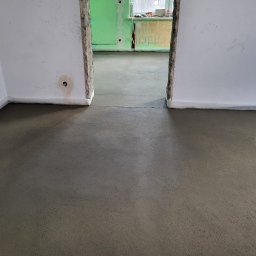 ANDROBUD - Wylana, świeża wylewka podłogowa w jasnym pomieszczeniu z widocznymi fragmentami ścian i przejściem do kolejnego pokoju w trakcie prac wykończeniowych.