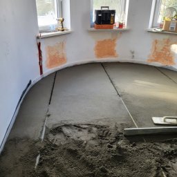 ANDROBUD - Wyrównywanie wylewki w zaokrąglonym pomieszczeniu. Widoczne narzędzia i częściowo wykończone ściany z oknami. Prace wykończeniowe w trakcie realizacji.