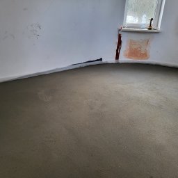 ANDROBUD - Wylana, świeża wylewka podłogowa w pomieszczeniu z przygotowanymi ścianami do dalszych prac wykończeniowych. Widoczne okna i narzędzia na parapecie.