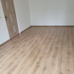 ANDROBUD - Jasne panele podłogowe ułożone w pustym pokoju z białą ścianą i drewnianymi drzwiami z przeszkleniami. Widok na podłogę z perspektywy.