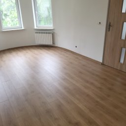 ANDROBUD - Jasne panele podłogowe w nowym pokoju z białymi ścianami, grzejnikiem i oknami. Widoczne drzwi z drewnianą okleiną. Świeżo wykończone wnętrze.