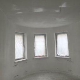 ANDROBUD - Wnętrze po szpachlowaniu: zaokrąglone ściany, trzy okna zabezpieczone folią, widoczne ślady po instalacji elektrycznej. Minimalistyczne wykończenie w odcieniach bieli.