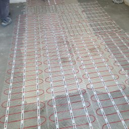 ANDROBUD - Rozłożona mata grzewcza Warmtec z czerwonymi przewodami na szarej posadzce w trakcie montażu ogrzewania podłogowego. Widoczne fragmenty nóg instalatora.