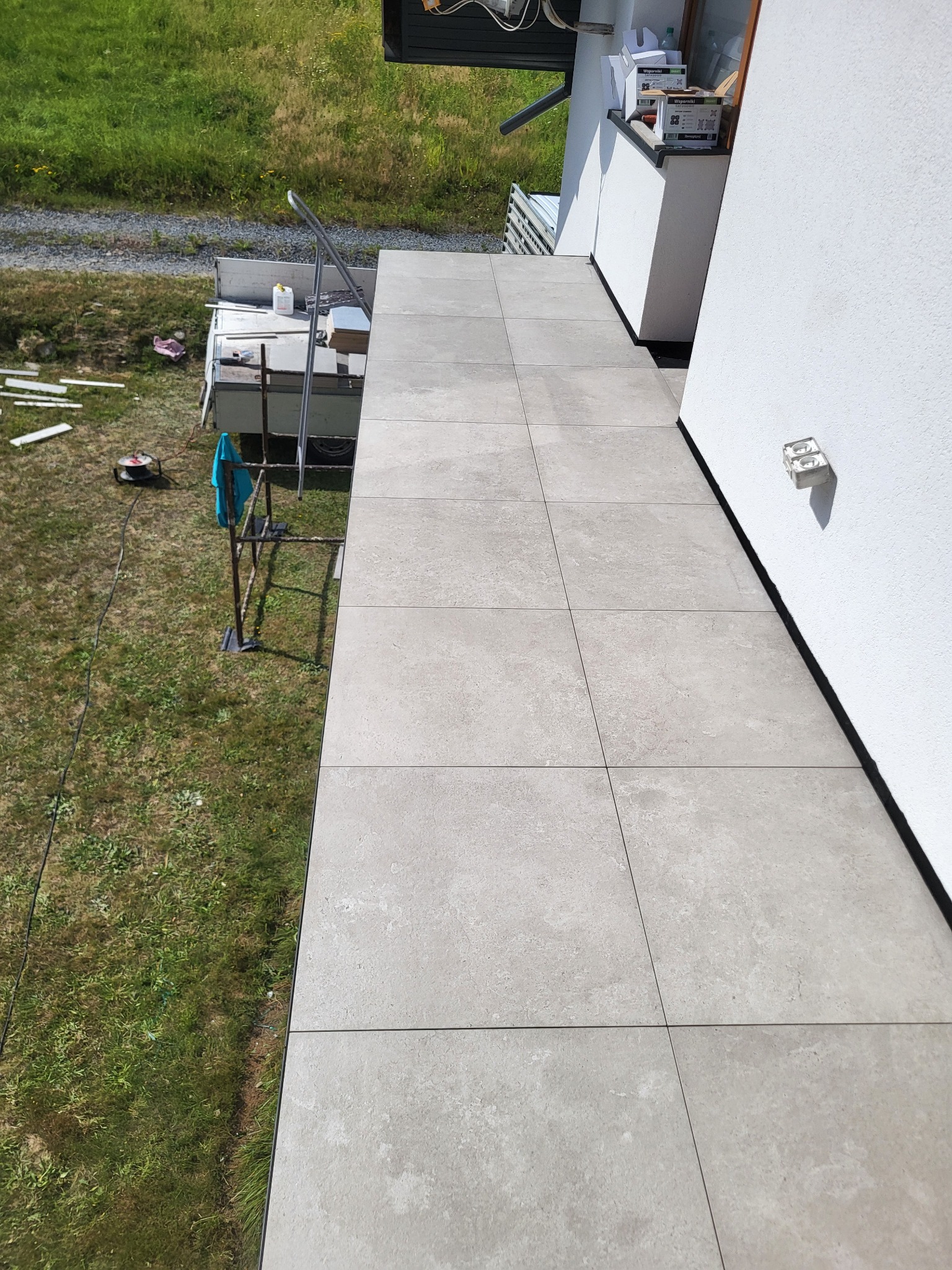 Wykończony taras z szarych, kwadratowych płytek, widok z góry. Na trawie widoczne narzędzia i materiały budowlane. Jasna elewacja budynku w tle.