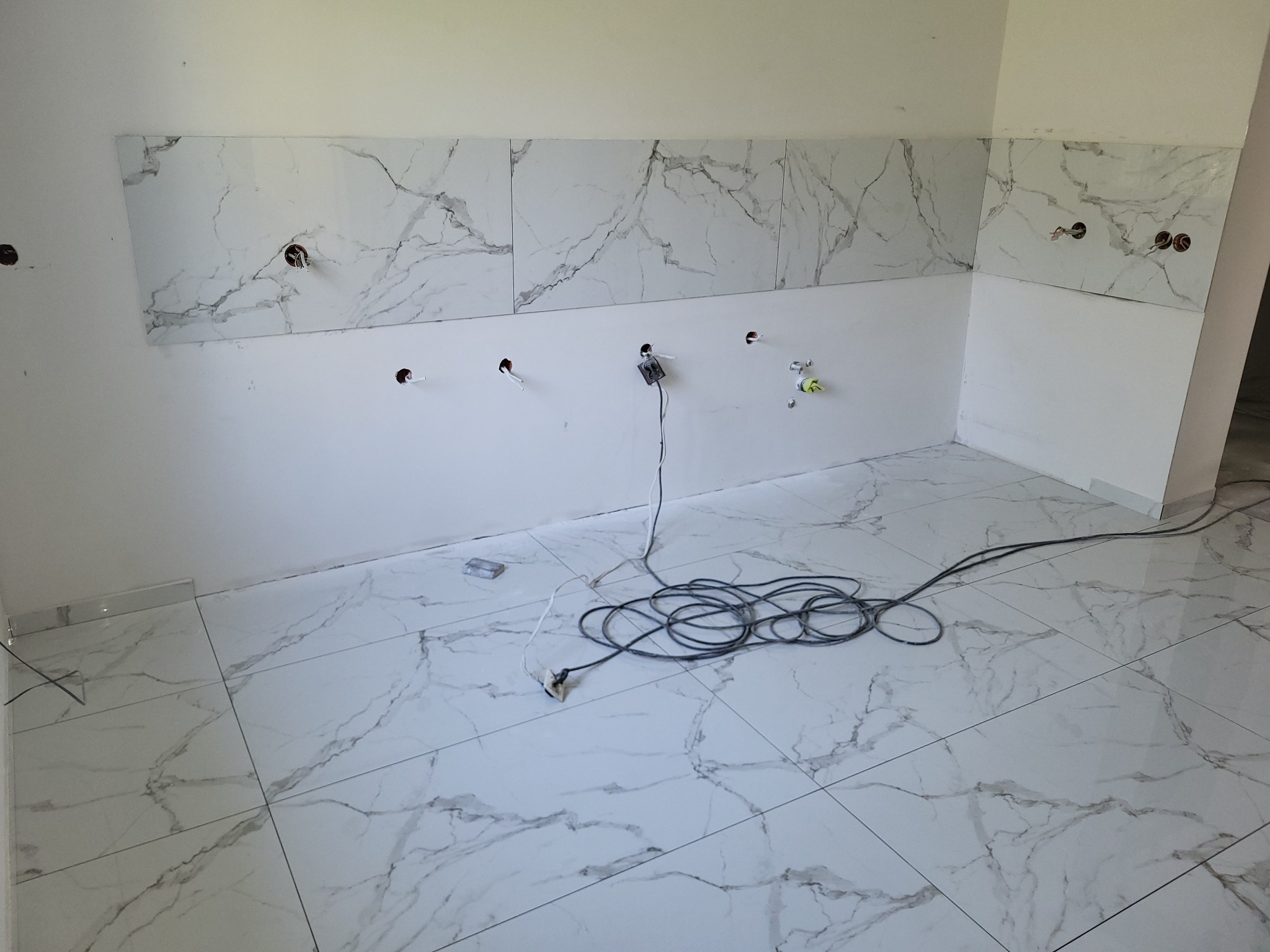 Białe płytki imitujące marmur na podłodze i częściowo na ścianie łazienki w trakcie montażu. Widoczne kable i otwory na instalacje.