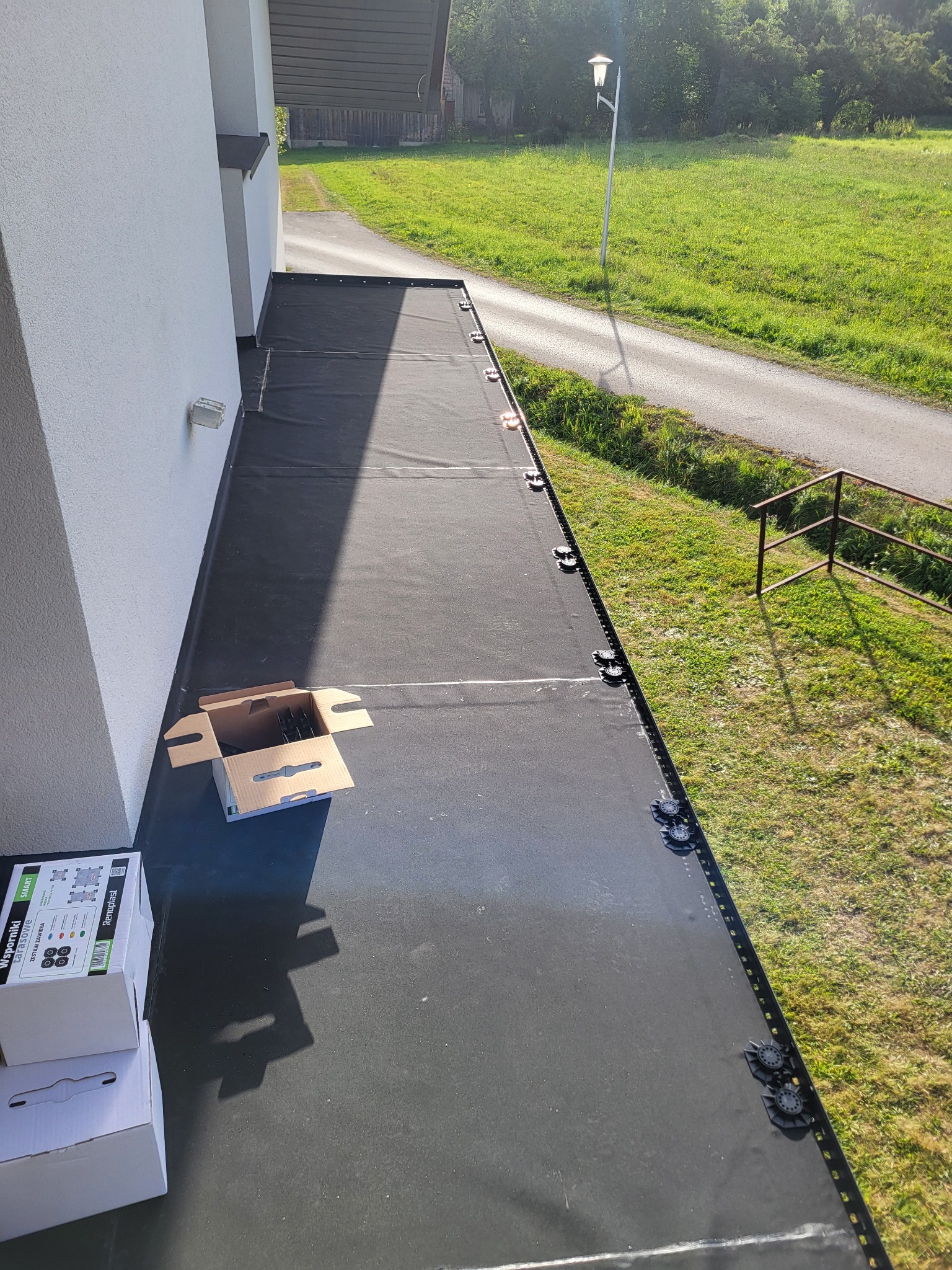 Czarna hydroizolacja na balkonie z widocznymi wspornikami tarasowymi i kartonami. Widok z góry na gotową powierzchnię z elementami montażowymi. Tło: trawa i droga.