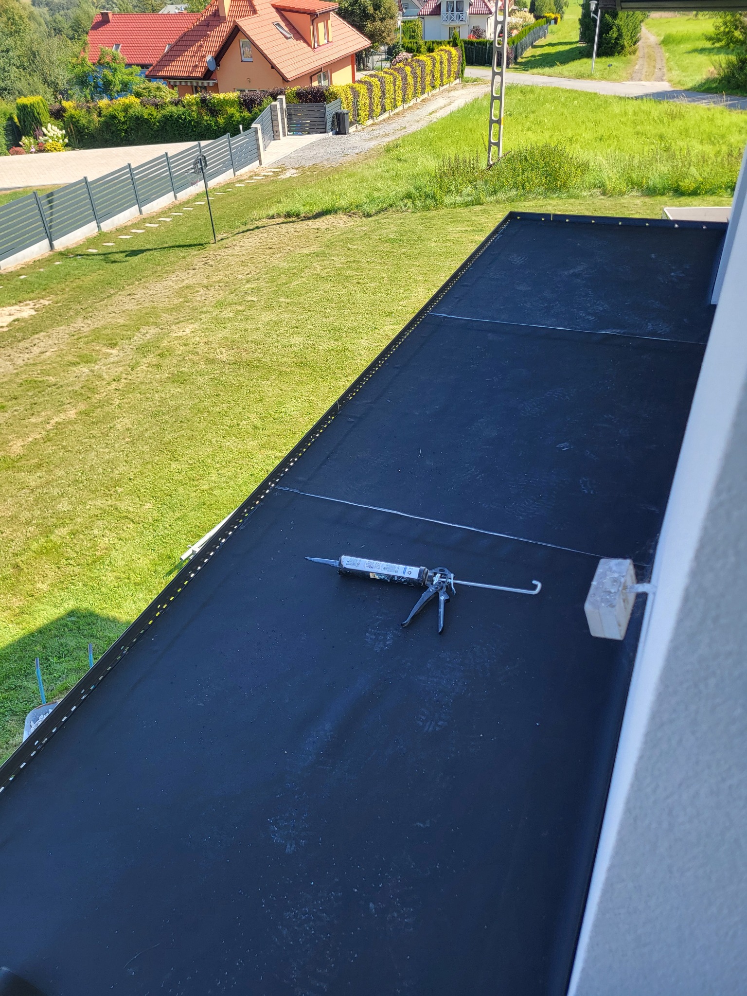 Czarna, gumowa membrana na dachu z narzędziem do uszczelniania, widok z góry na tle zieleni. Detal wykończenia dachu z bliska, z narzędziem do aplikacji uszczelniacza.