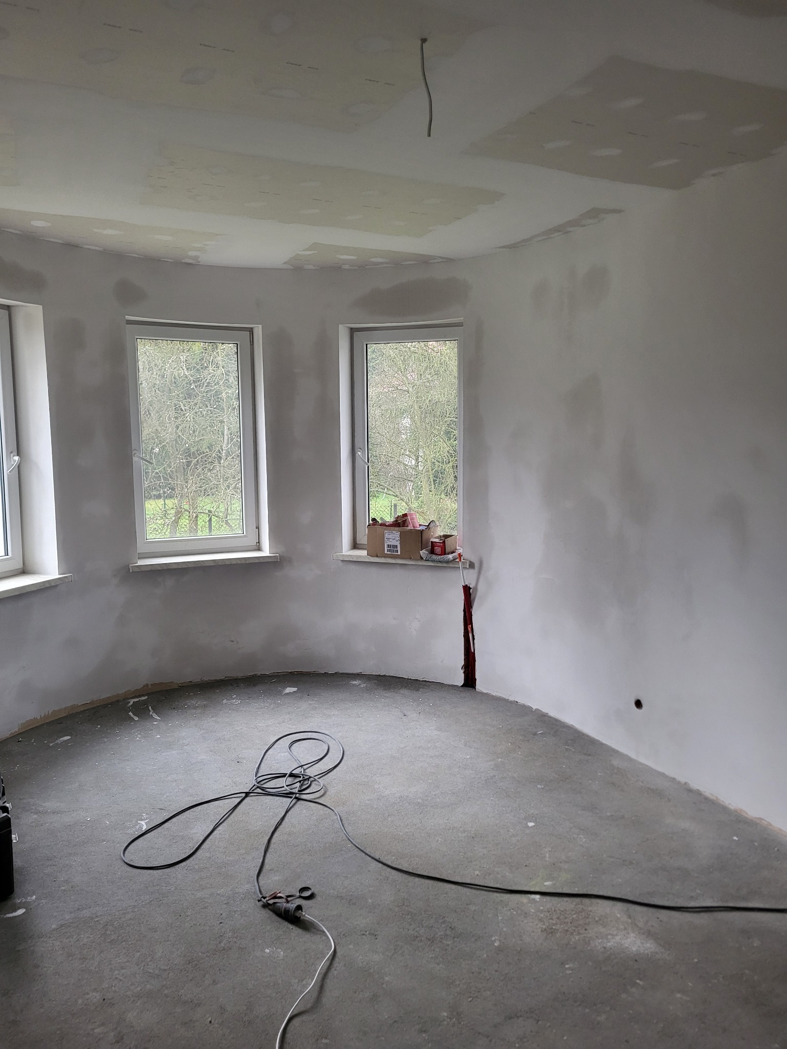 Wnętrze okrągłego pokoju w trakcie szpachlowania ścian i sufitu. Widoczne trzy okna, szpachlowane ściany, sufit z płyt kartonowo-gipsowych i kable na podłodze.