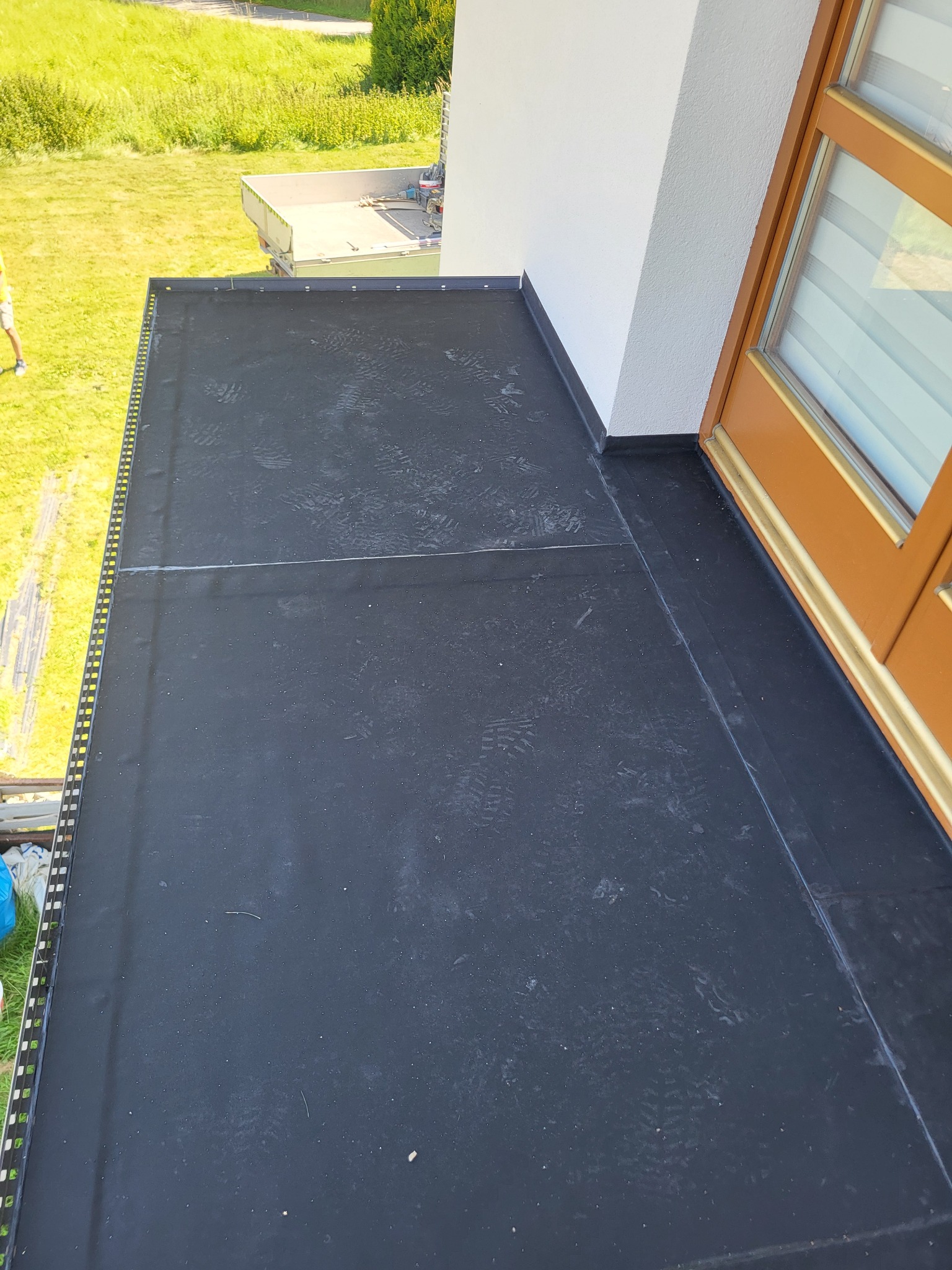 Wykończony balkon z czarną, hydroizolacyjną powłoką, widoczne ślady stóp. Detal wykończenia przy drzwiach balkonowych i metalowe obrzeże. Trawa w tle.