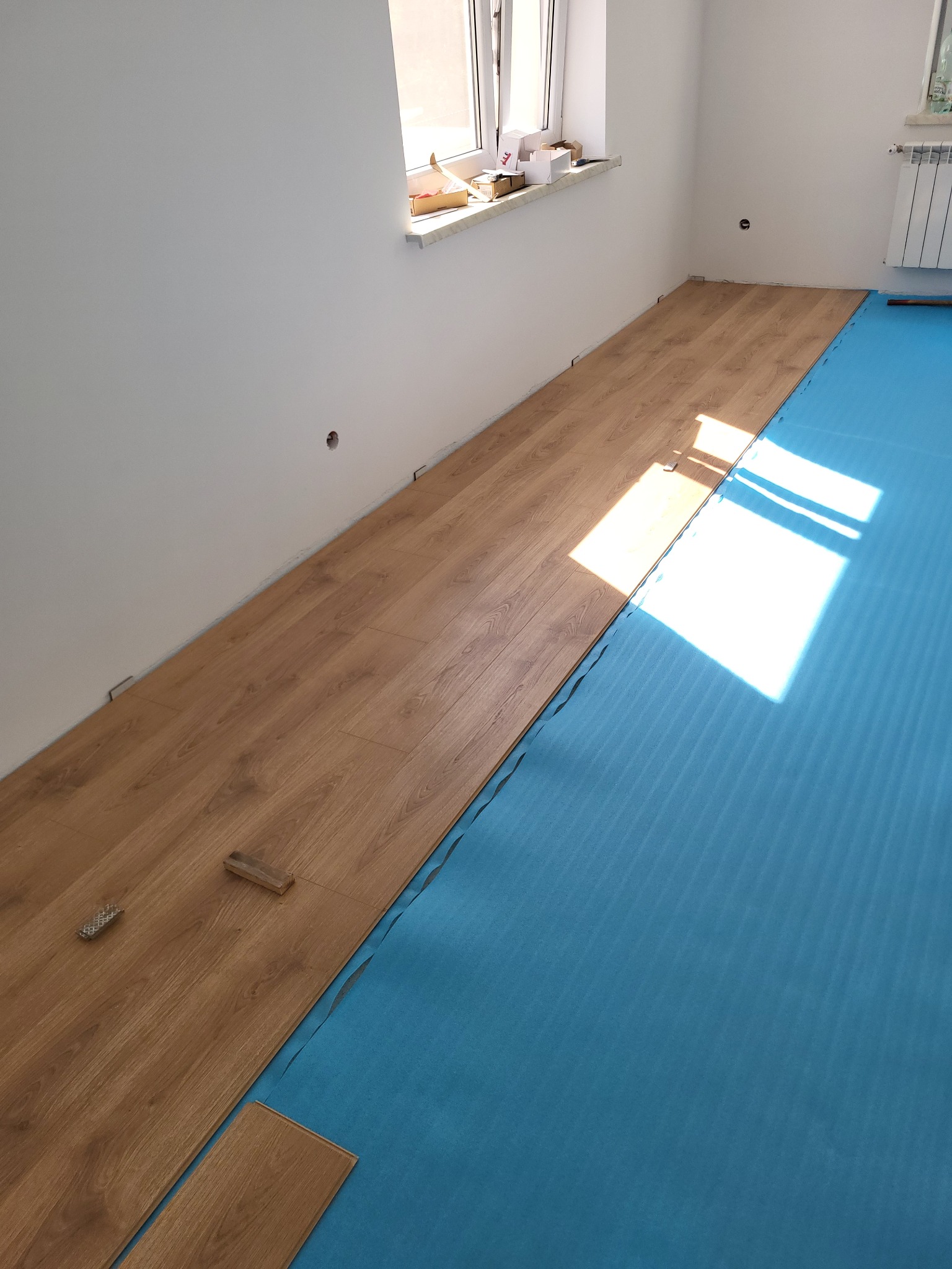 Układanie paneli podłogowych w jasnym wnętrzu. Częściowo ułożona podłoga z paneli w odcieniach drewna, podkład z niebieskiej pianki. Widoczne narzędzia i fragment okna.