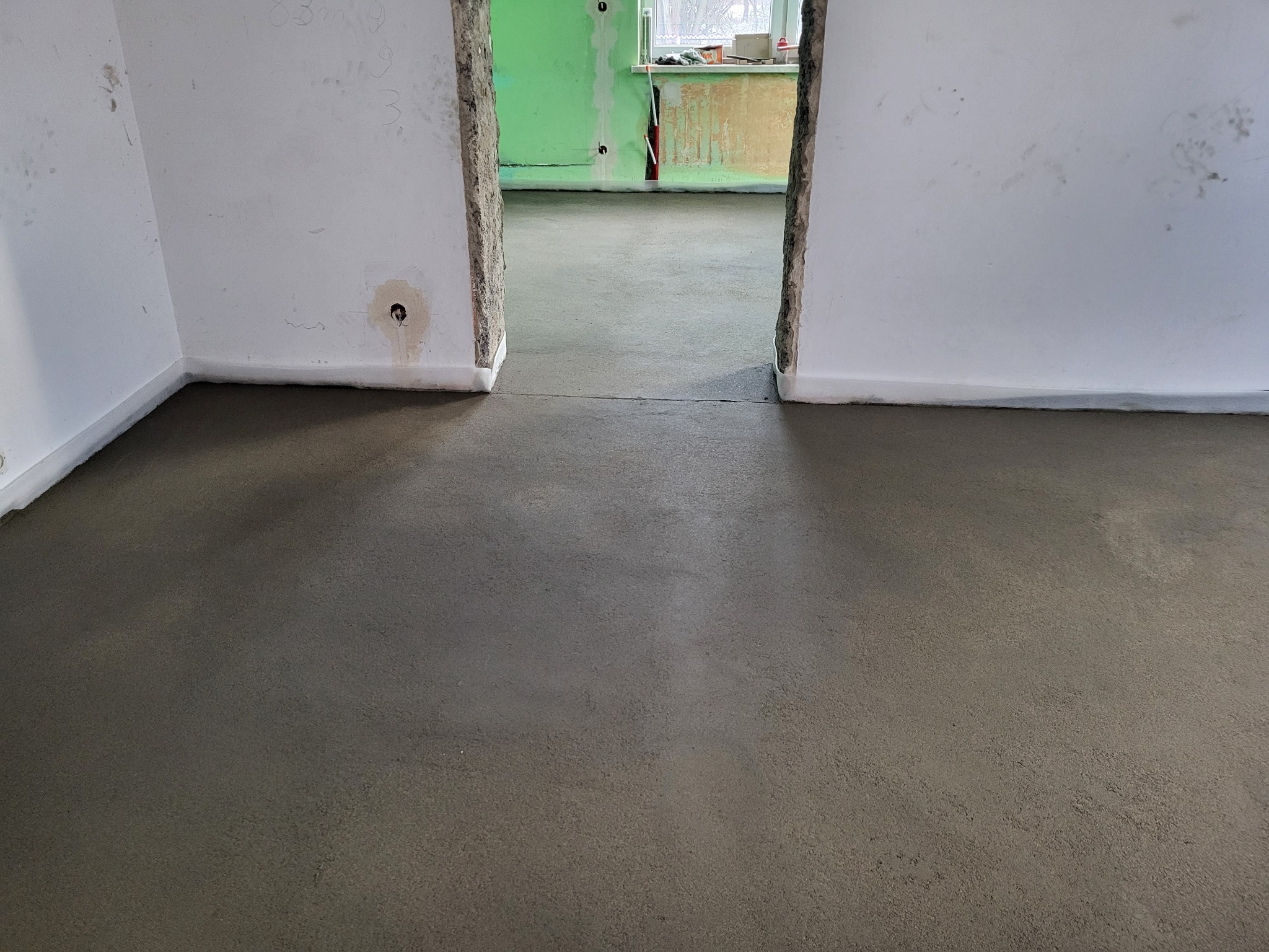 Wylana, świeża wylewka podłogowa w jasnym pomieszczeniu z widocznymi fragmentami ścian i przejściem do kolejnego pokoju w trakcie prac wykończeniowych.