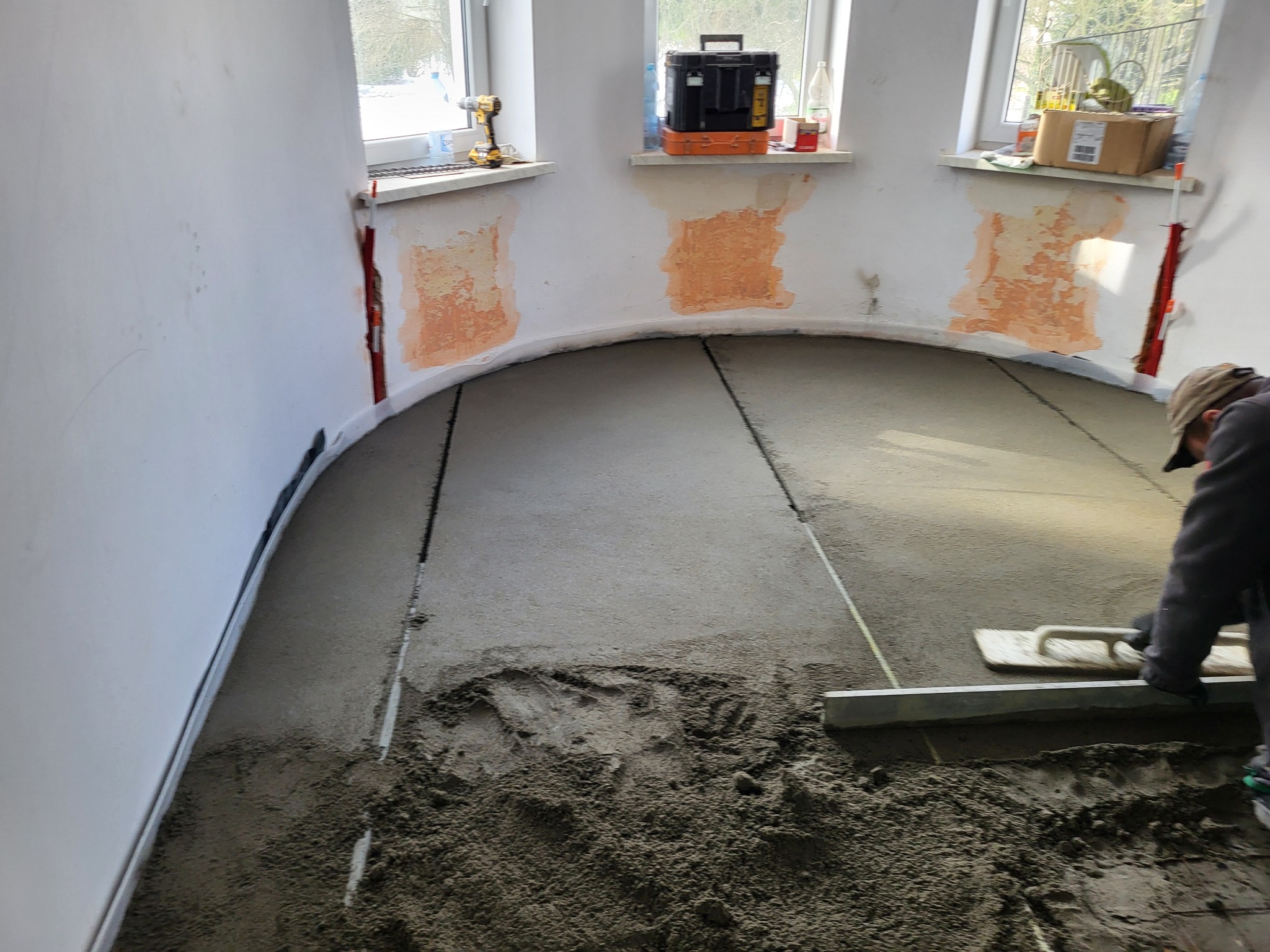 Wyrównywanie wylewki w zaokrąglonym pomieszczeniu. Widoczne narzędzia i częściowo wykończone ściany z oknami. Prace wykończeniowe w trakcie realizacji.