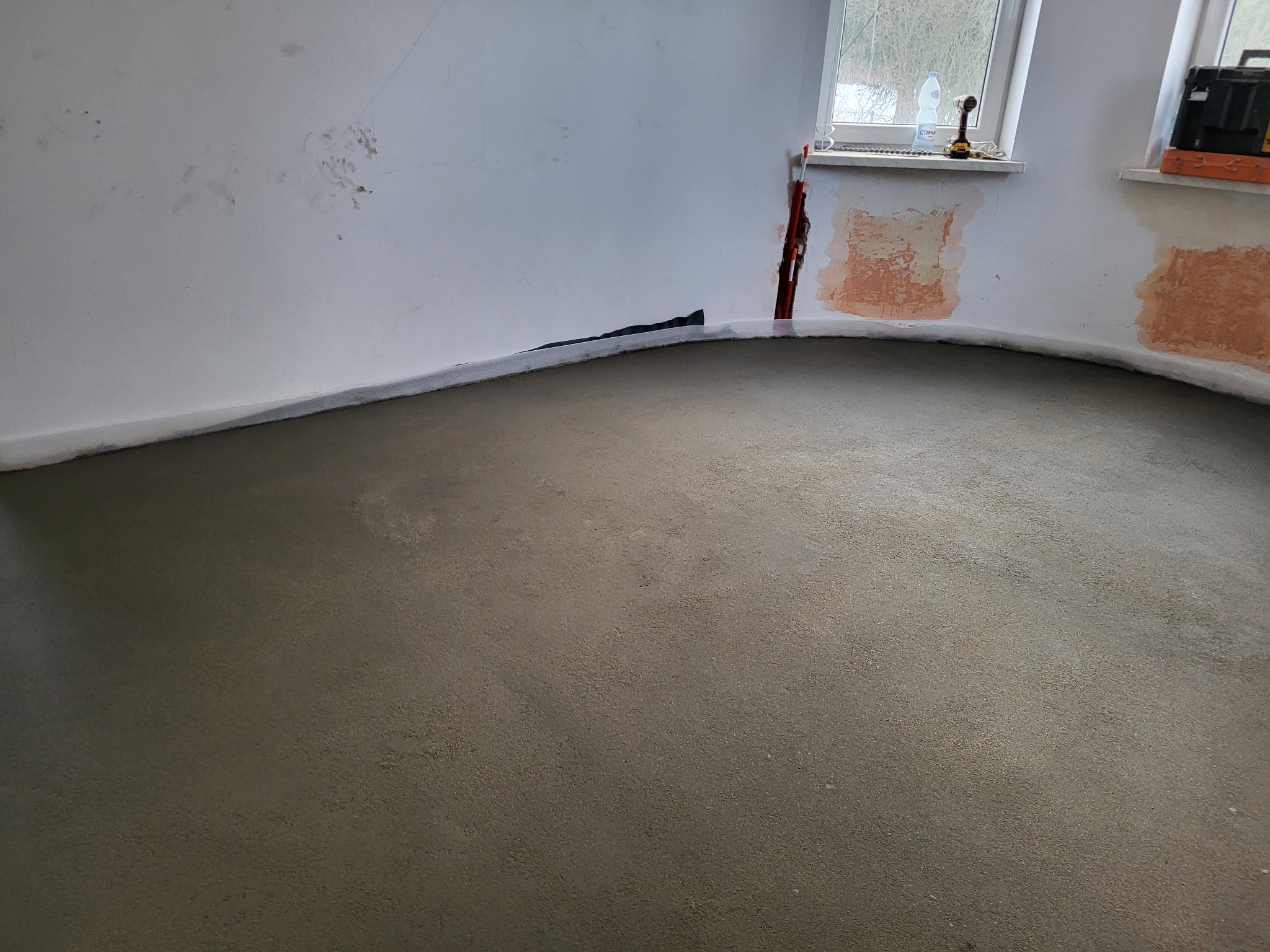 Wylana, świeża wylewka podłogowa w pomieszczeniu z przygotowanymi ścianami do dalszych prac wykończeniowych. Widoczne okna i narzędzia na parapecie.