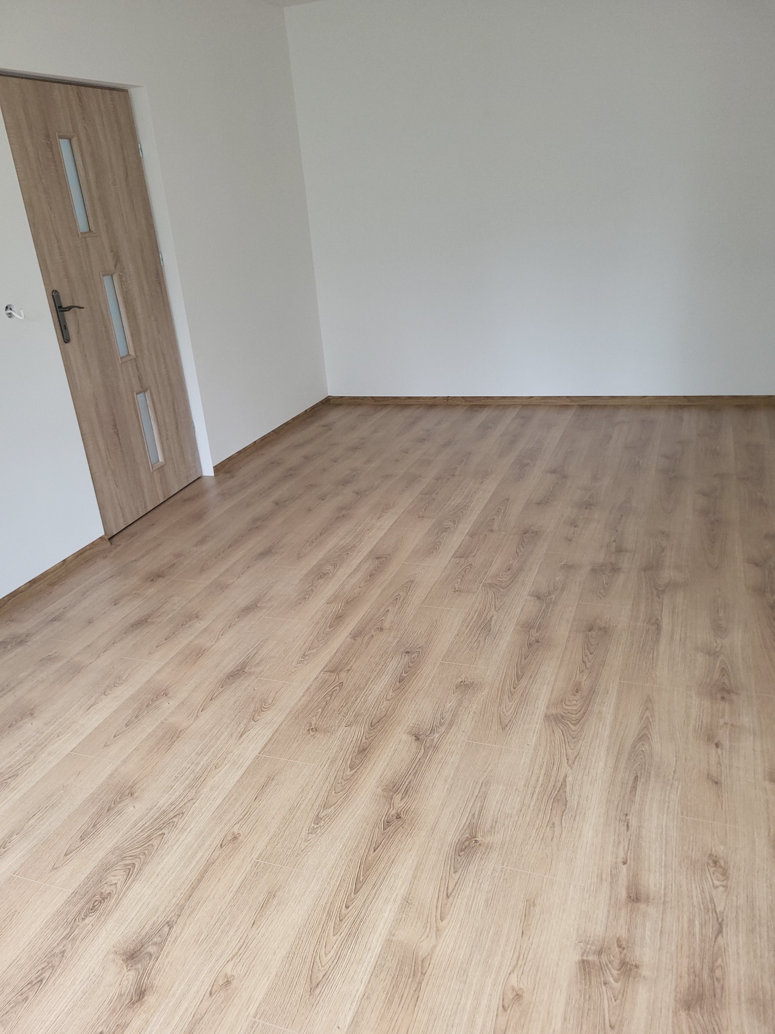 Jasne panele podłogowe ułożone w pustym pokoju z białą ścianą i drewnianymi drzwiami z przeszkleniami. Widok na podłogę z perspektywy.