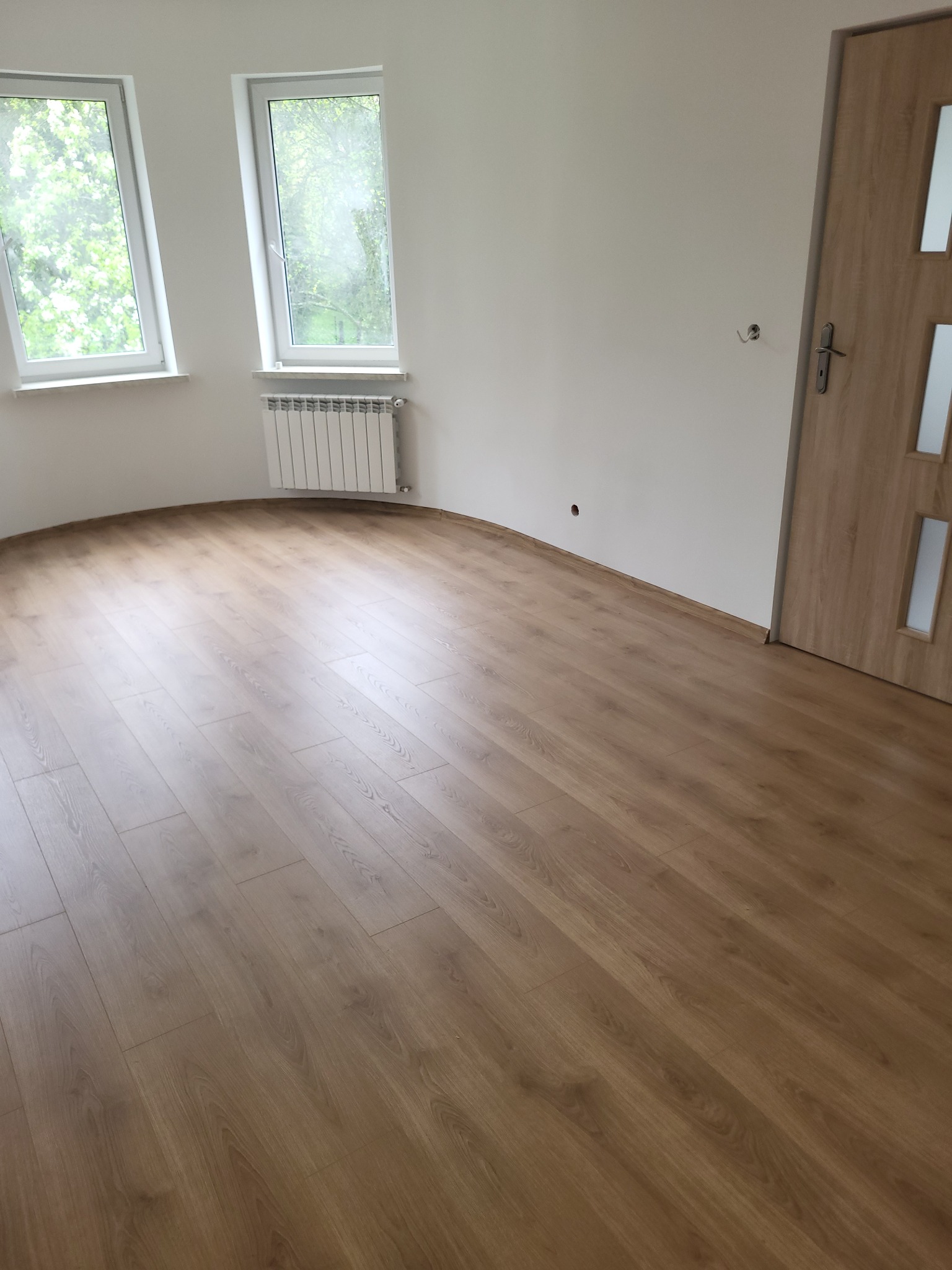 Jasne panele podłogowe w nowym pokoju z białymi ścianami, grzejnikiem i oknami. Widoczne drzwi z drewnianą okleiną. Świeżo wykończone wnętrze.