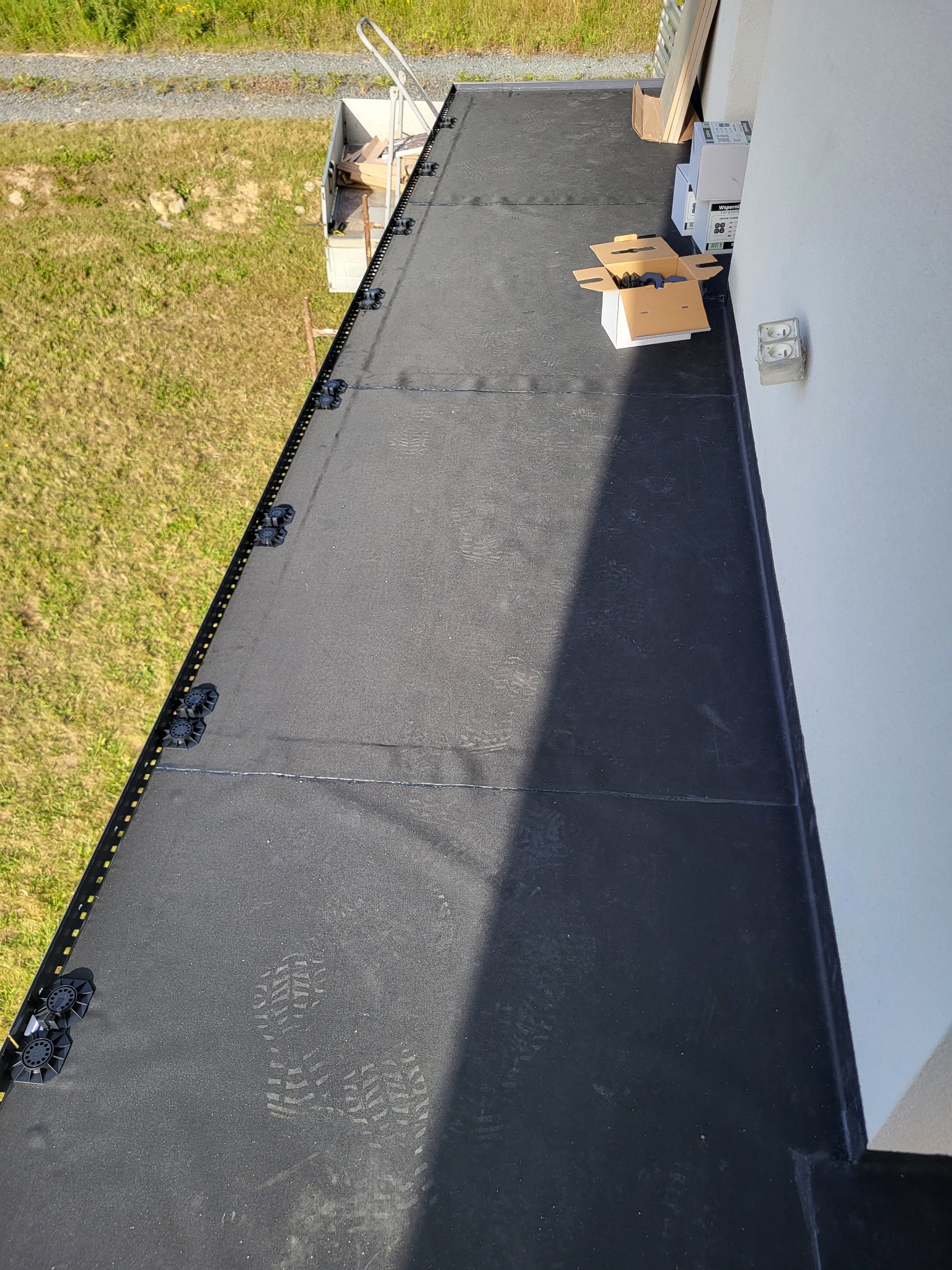 Czarna, płaska powierzchnia balkonu z widocznymi śladami butów, listwą przypodłogową i elementami montażowymi. W tle trawa i drabina.