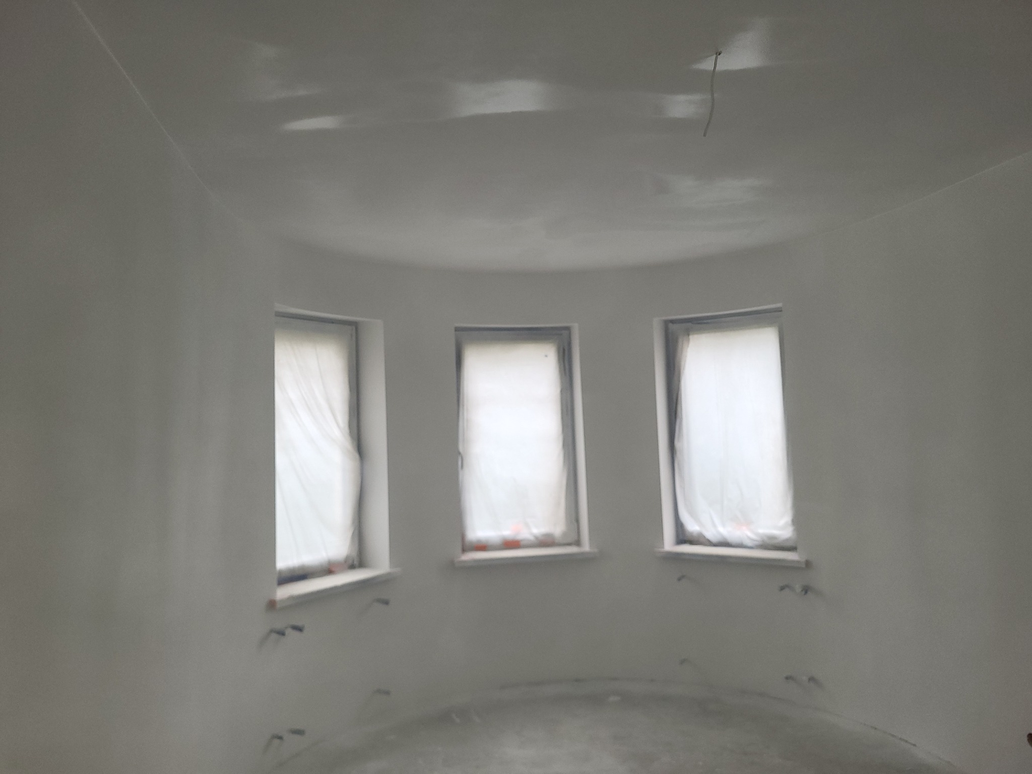 Wnętrze po szpachlowaniu: zaokrąglone ściany, trzy okna zabezpieczone folią, widoczne ślady po instalacji elektrycznej. Minimalistyczne wykończenie w odcieniach bieli.
