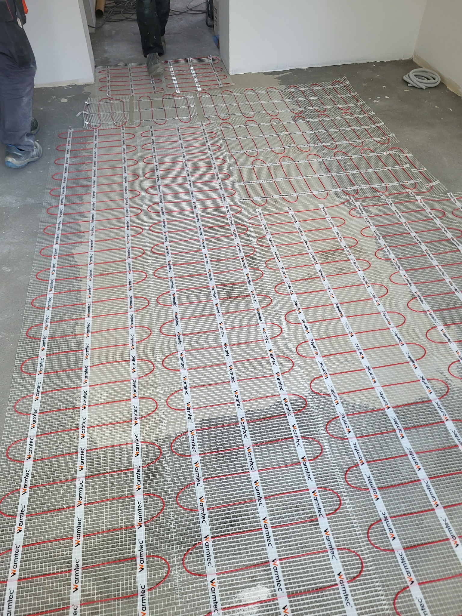 Rozłożona mata grzewcza Warmtec z czerwonymi przewodami na szarej posadzce w trakcie montażu ogrzewania podłogowego. Widoczne fragmenty nóg instalatora.