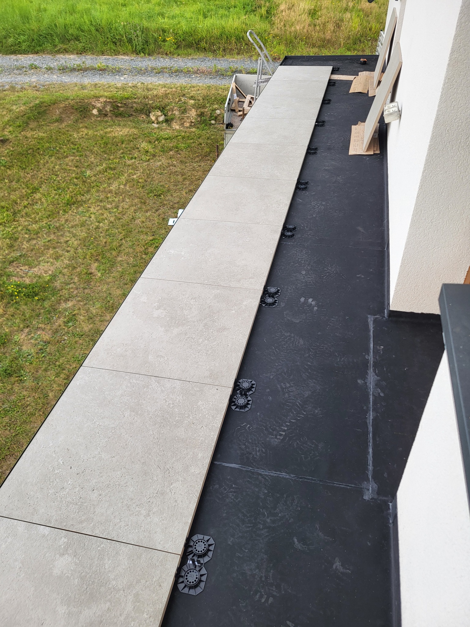 Montaż jasnych płyt na wspornikach na czarnej hydroizolacji balkonu, widok z góry. Częściowo ułożona podłoga balkonowa z widocznymi elementami montażowymi.