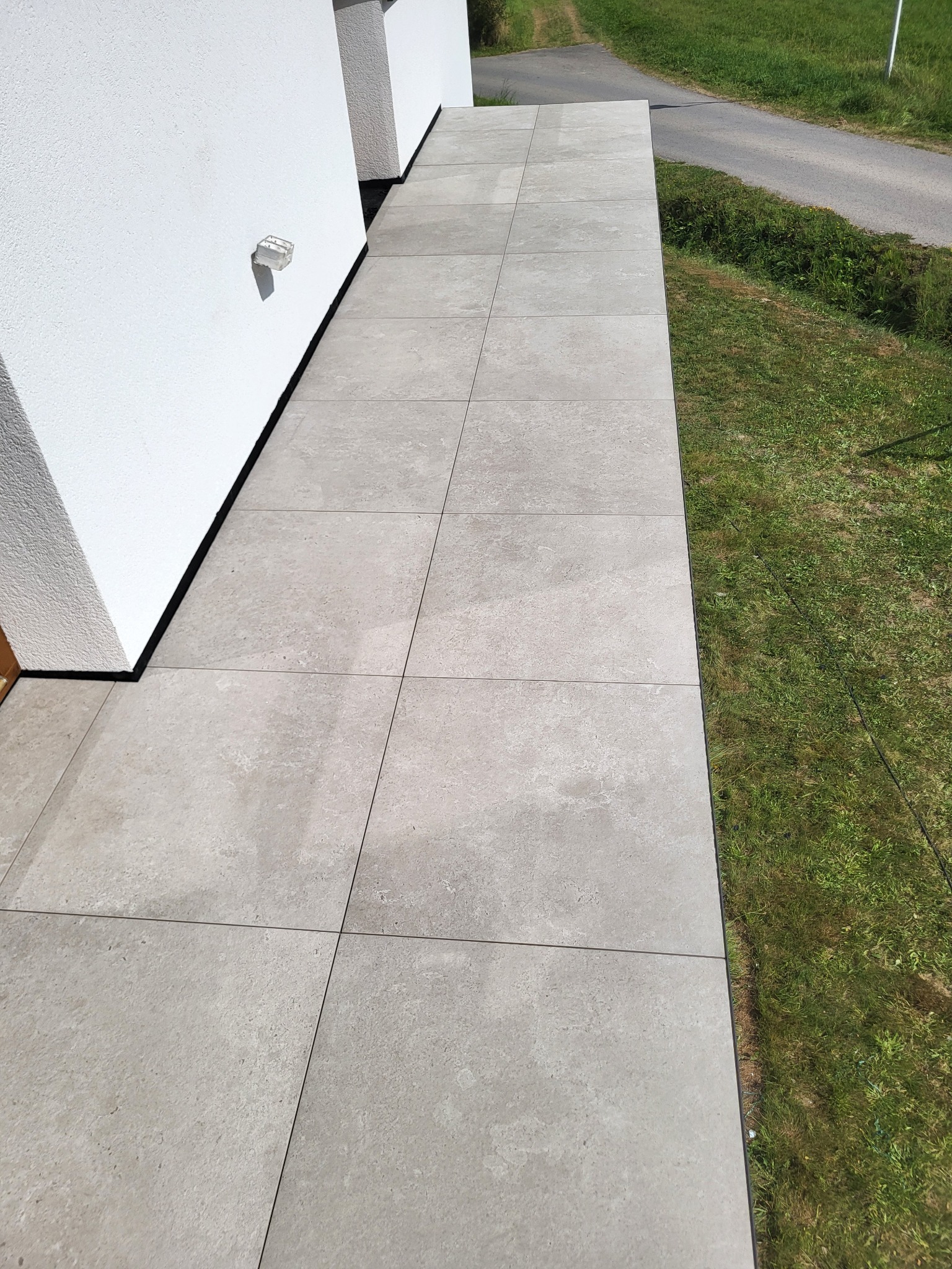Jasnoszara, gładka posadzka z płytek na zewnątrz budynku, przylegająca do białej ściany. Widok z góry na fragment trawnika i drogę w tle. Minimalistyczny design.