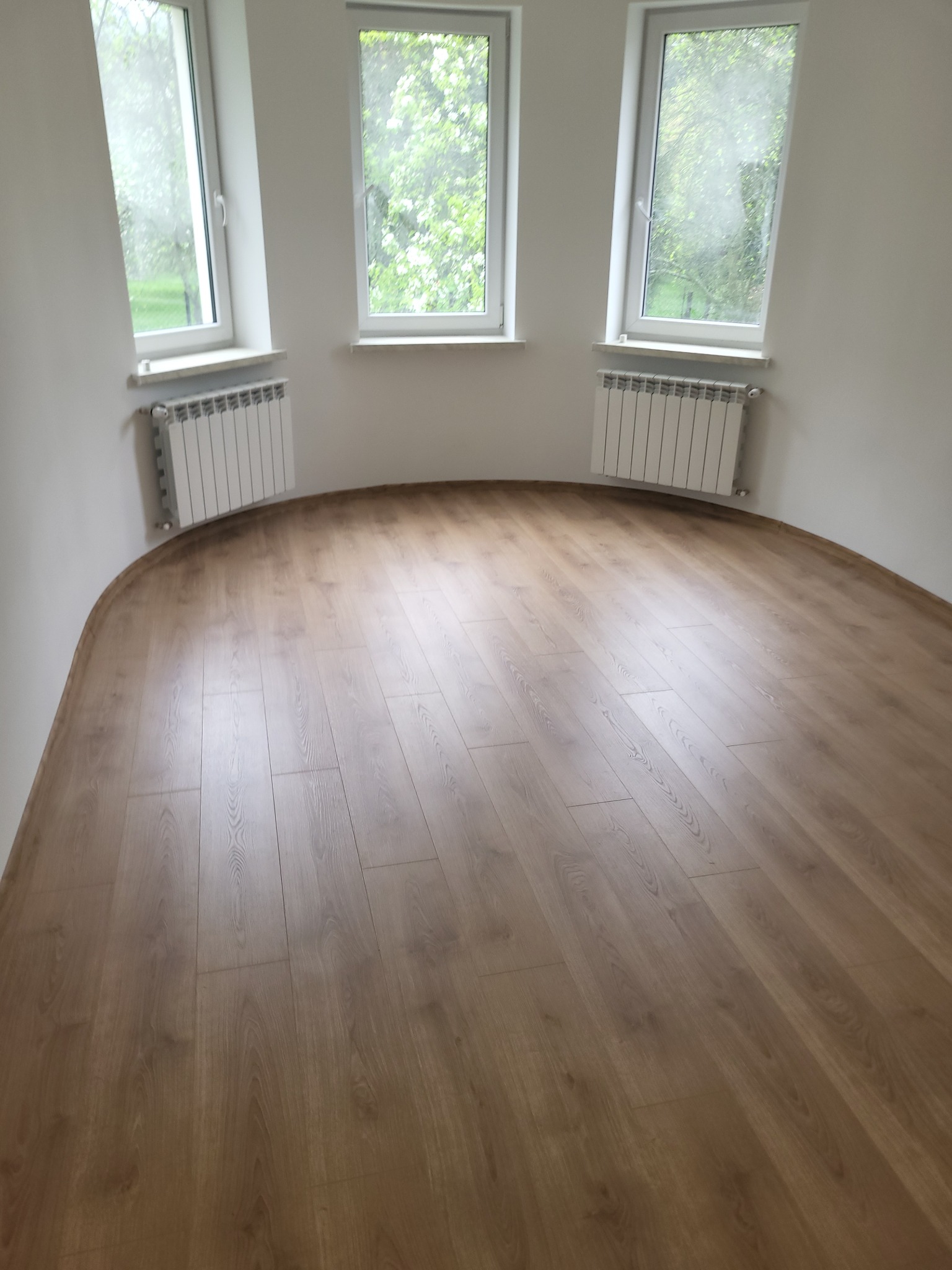 Jasne panele podłogowe ułożone w pokoju z trzema oknami i białymi grzejnikami. Widok z góry, ukazujący estetyczne wykończenie podłogi.