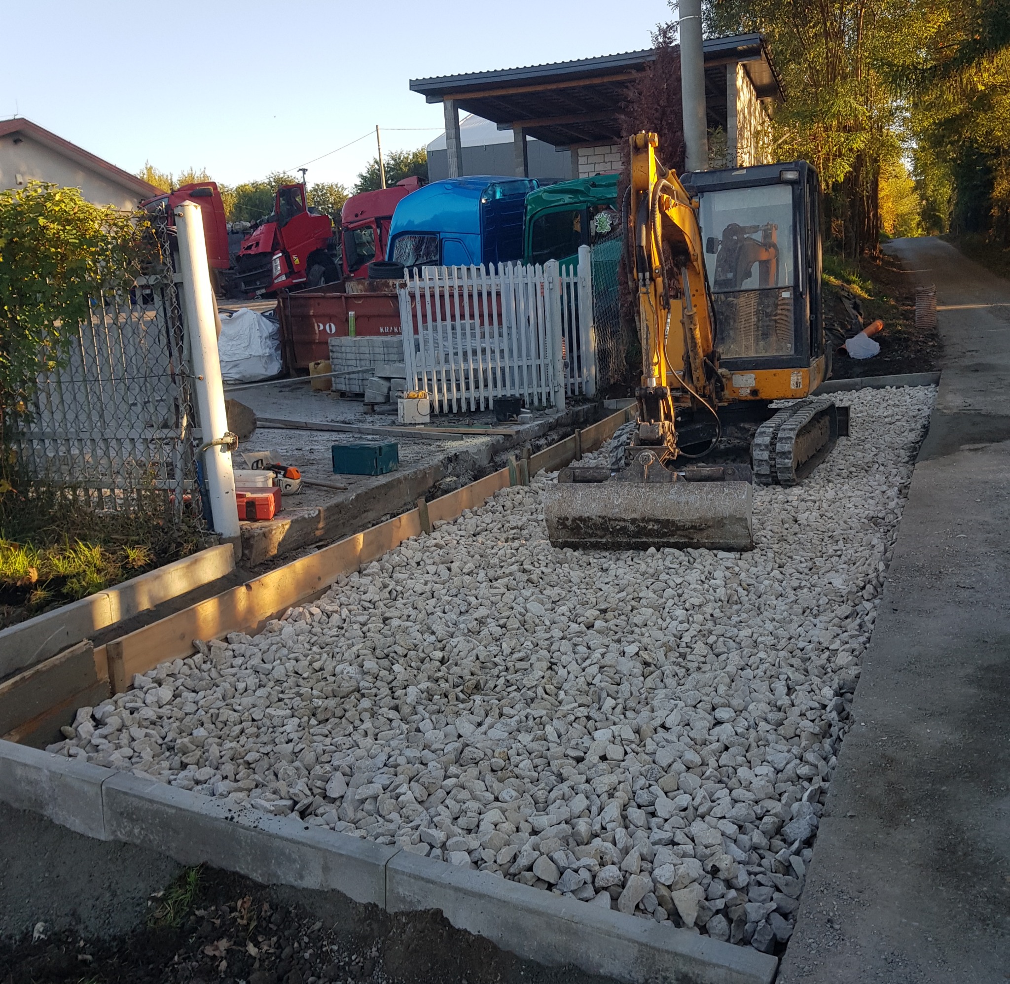 Żółta minikoparka na placu budowy, wysypany tłuczeń, obrzeża betonowe i drewniane szalunki, w tle zaparkowane ciężarówki.