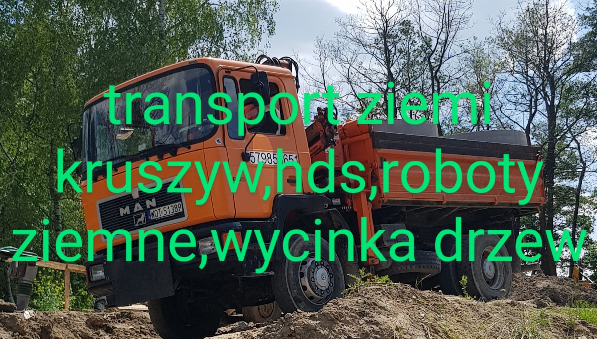 Pomarańczowa ciężarówka MAN na nierównym, piaszczystym terenie z widoczną ziemią, drzewami w tle i ubijarką do gruntu w pobliżu, sugerująca prace ziemne.