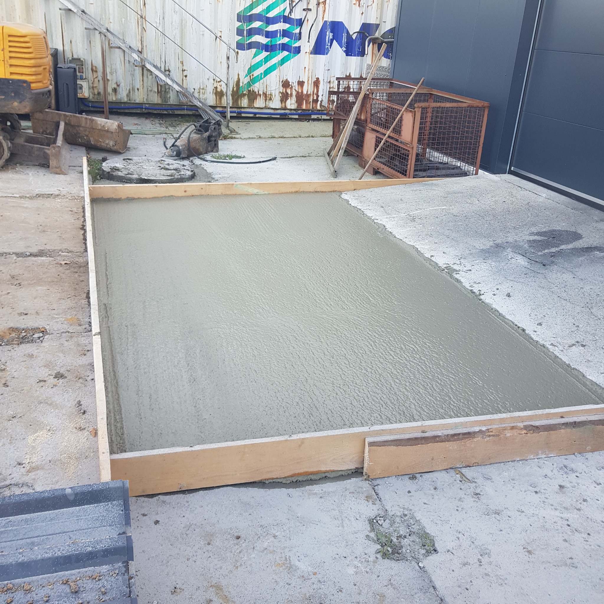 Świeżo wylany beton w drewnianym szalunku, obok stoi żółta mini koparka i metalowe klatki, w tle widoczny magazyn z logo.