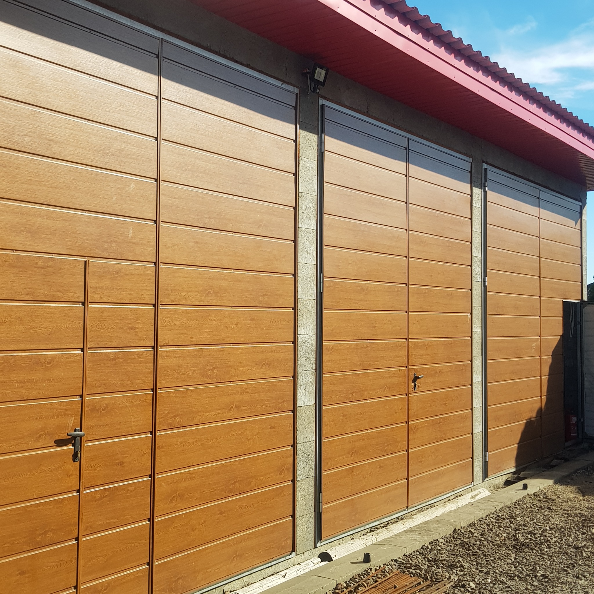 Trzy brązowe, segmentowe bramy garażowe z imitacją drewna, oddzielone betonowymi filarami, pod czerwoną konstrukcją dachu, na tle jasnego nieba.