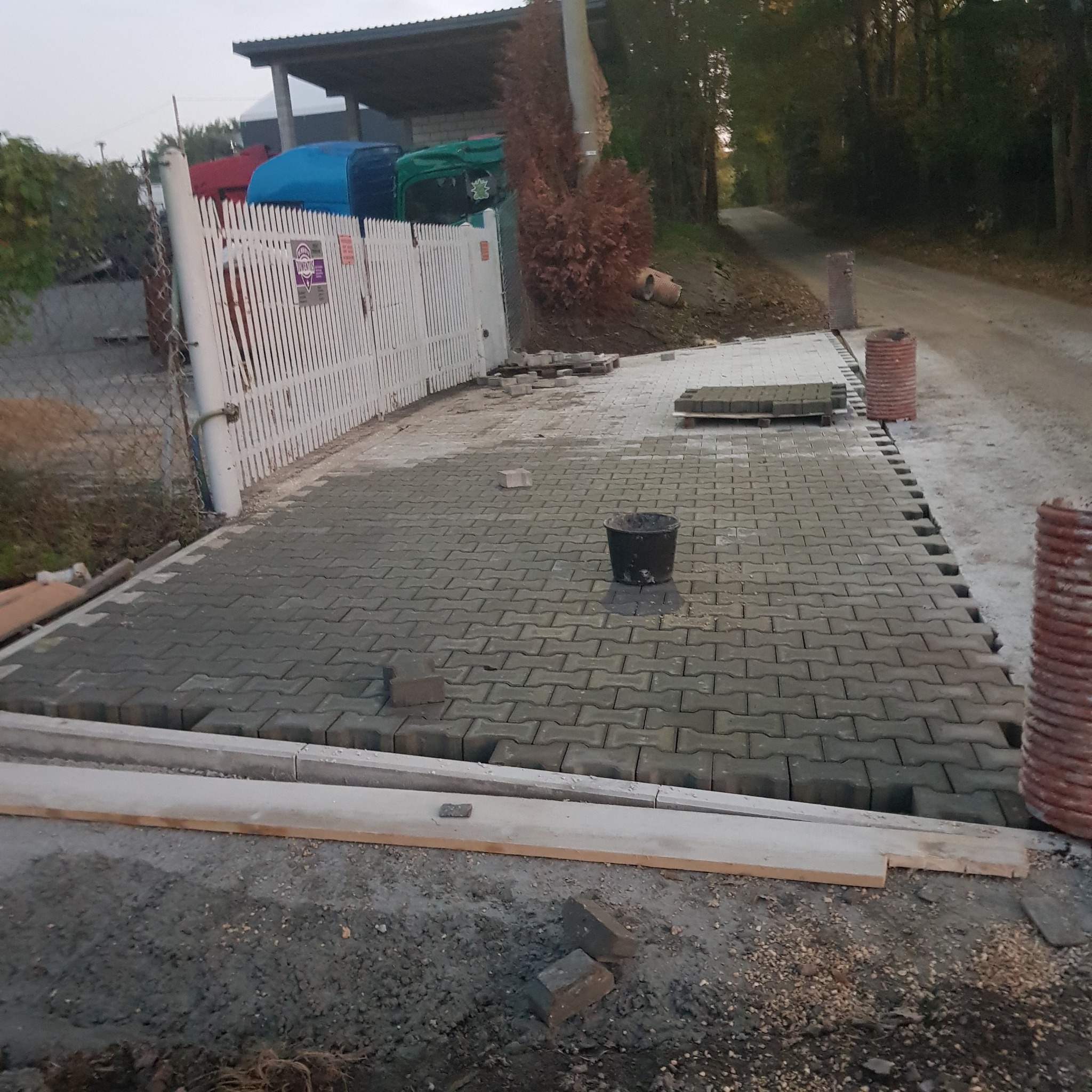 Układanie szarej kostki brukowej na podjeździe, widoczne obrzeża, czarne wiadro, palety z kostką i fragment drogi gruntowej w tle.