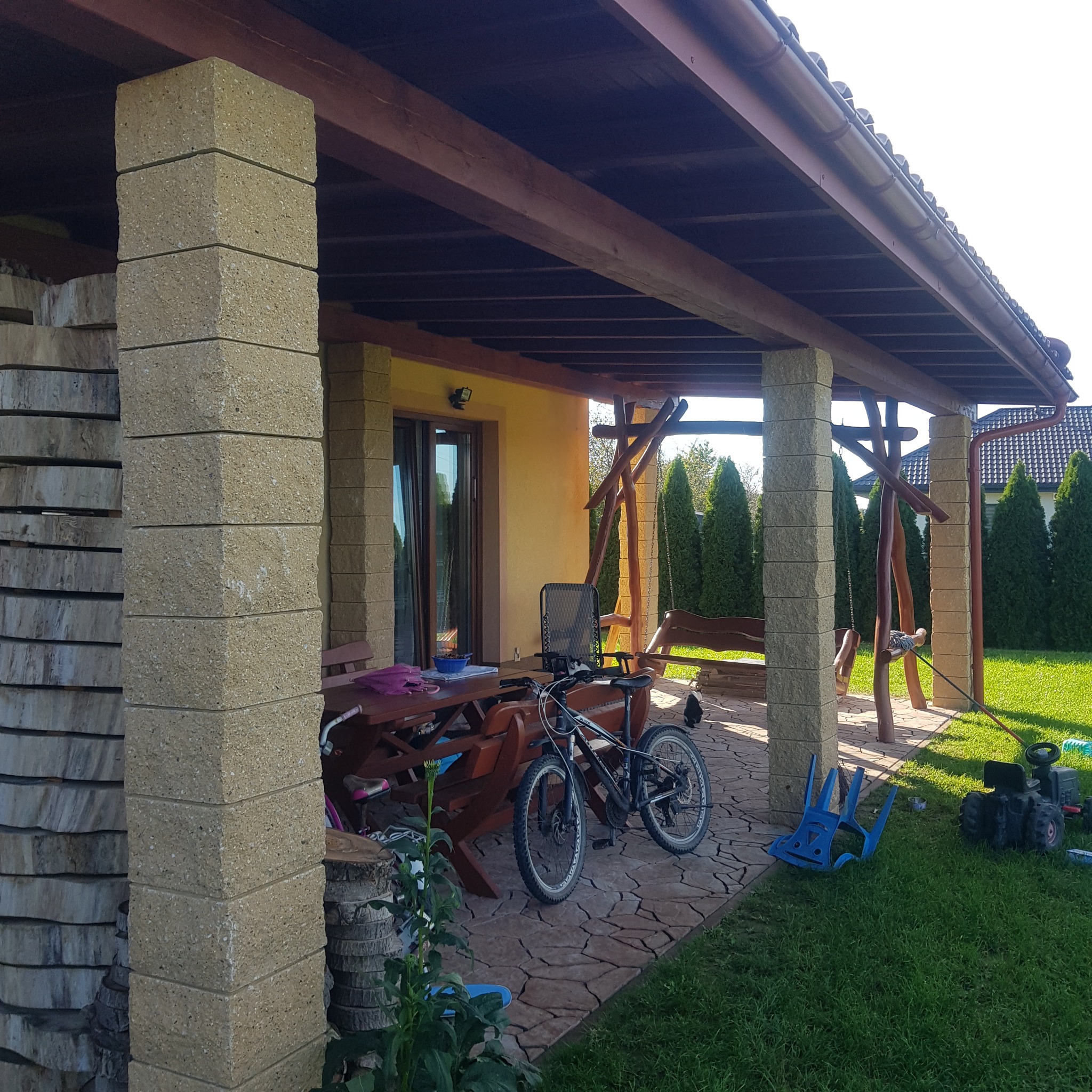 Zadaszony taras z kamienną podłogą i filarami, drewniana huśtawka, rower oparty o stół, trawnik z kosiarką-zabawką w tle.