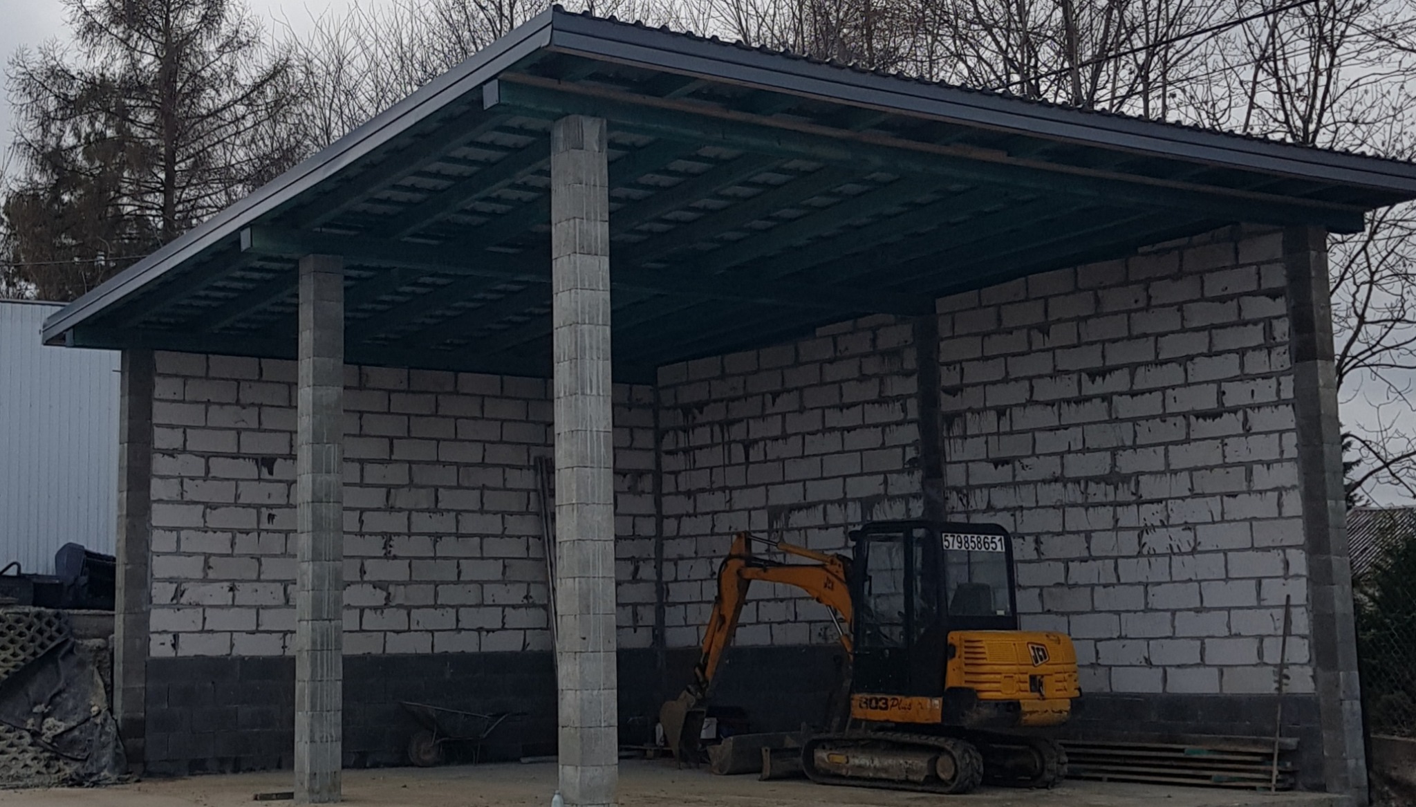 Niedokończona wiata z pustaków betonowych z żółtą mini koparką JCB 803 Plus w środku, widoczne betonowe słupy i drewniana konstrukcja dachu.