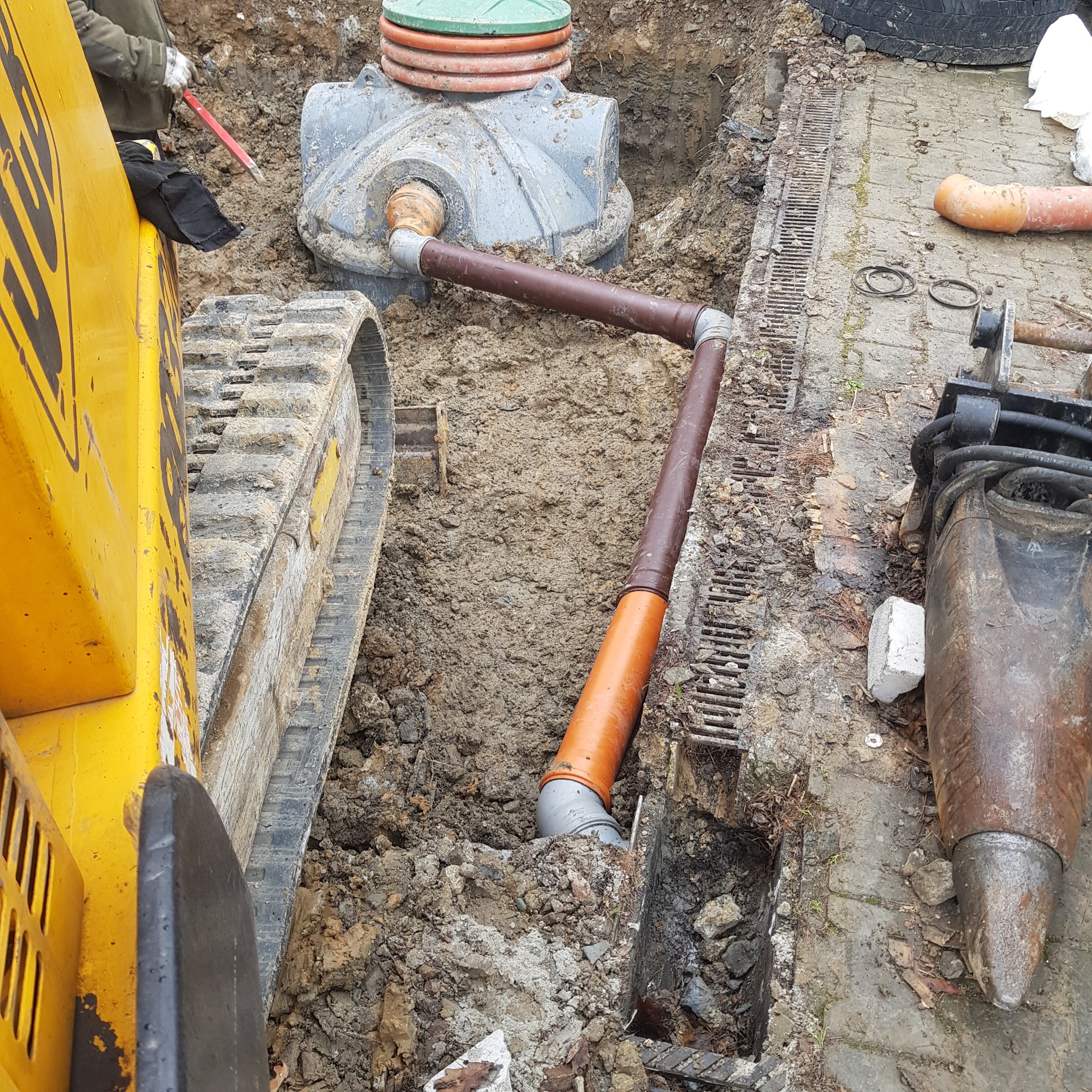 Wykonanie przyłącza kanalizacyjnego z wykorzystaniem koparki gąsienicowej, widoczne rury kanalizacyjne, studzienka rewizyjna z zieloną pokrywą oraz koryto odwadniające.