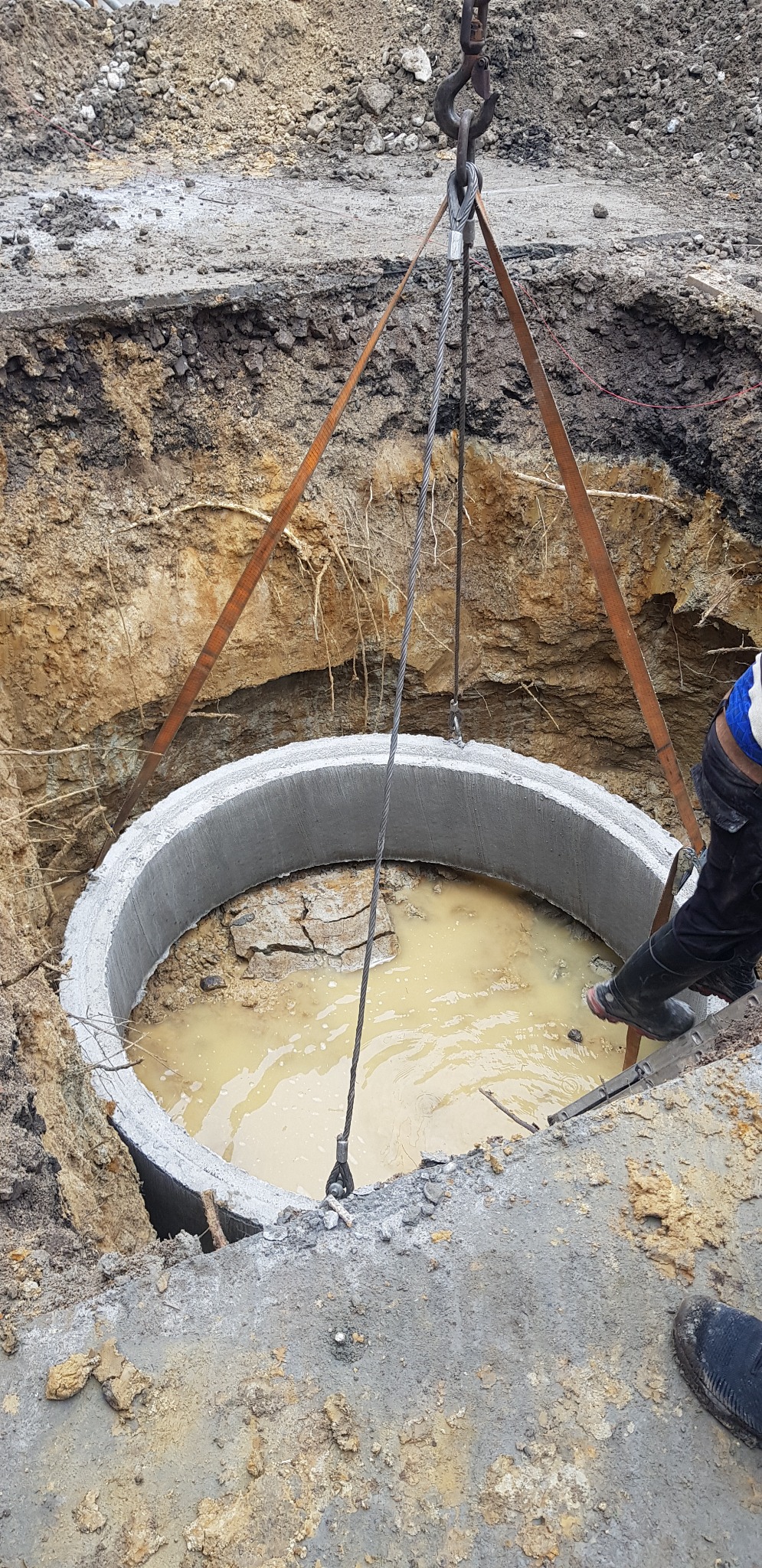Betonowy krąg studni opuszczany do wykopu za pomocą dźwigu linowego, wewnątrz woda zabarwiona ziemią, widoczne ślady pracy ziemnej i fragment buta pracownika.