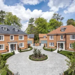 Dwa identyczne, dwupiętrowe domy z czerwonej cegły z szarymi dachami, symetrycznie usytuowane po obu stronach okrągłego podjazdu z kostki brukowej, otoczone zielenią i drzewami.